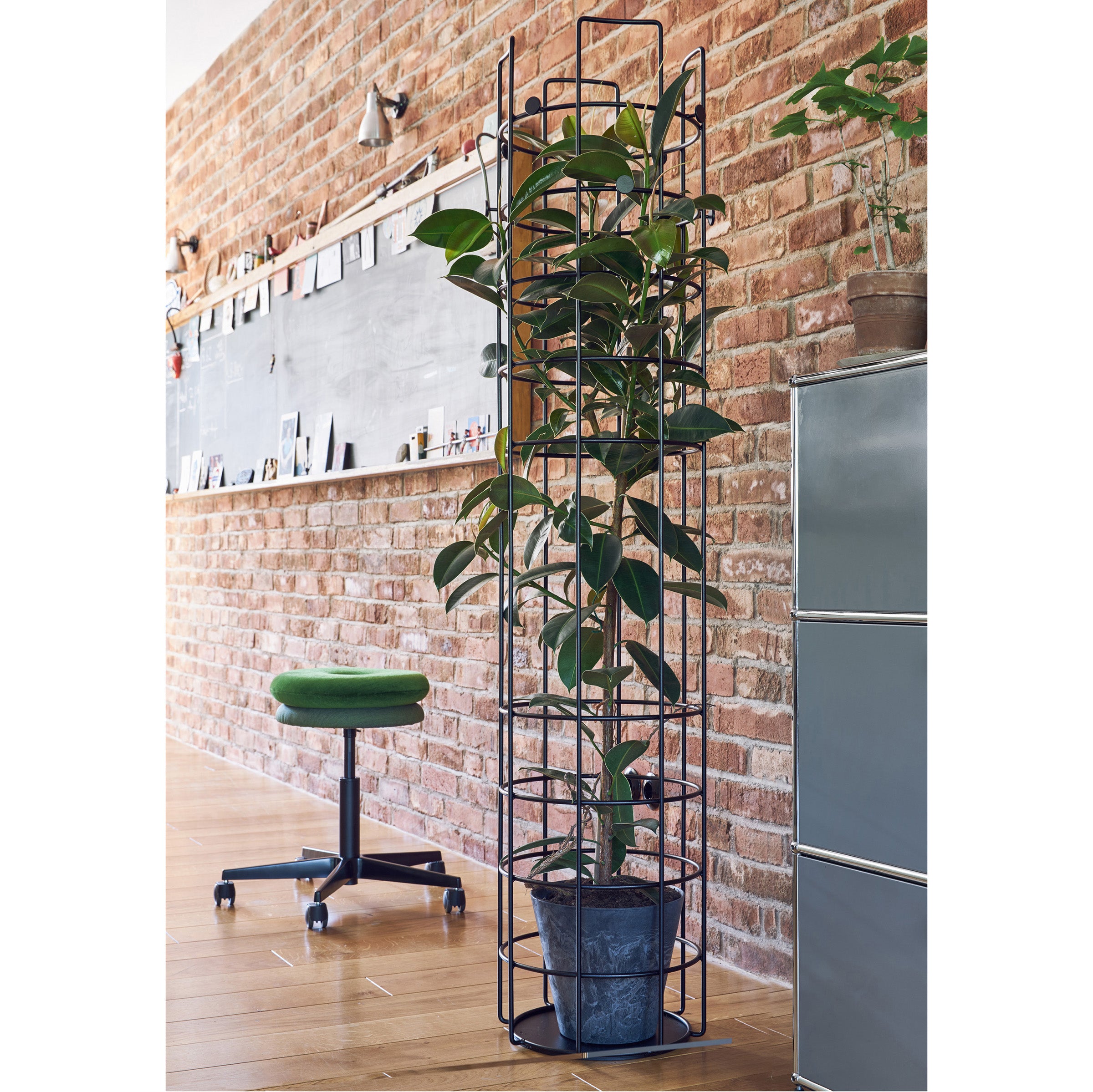 A Bazar Coat Stand by Richard Lampert, holding a potted plant, stands in front of a brick wall. A blackboard with pinned items hangs on the wall, and nearby, a green wheeled stool and gray filing cabinet sit on the wooden floor.