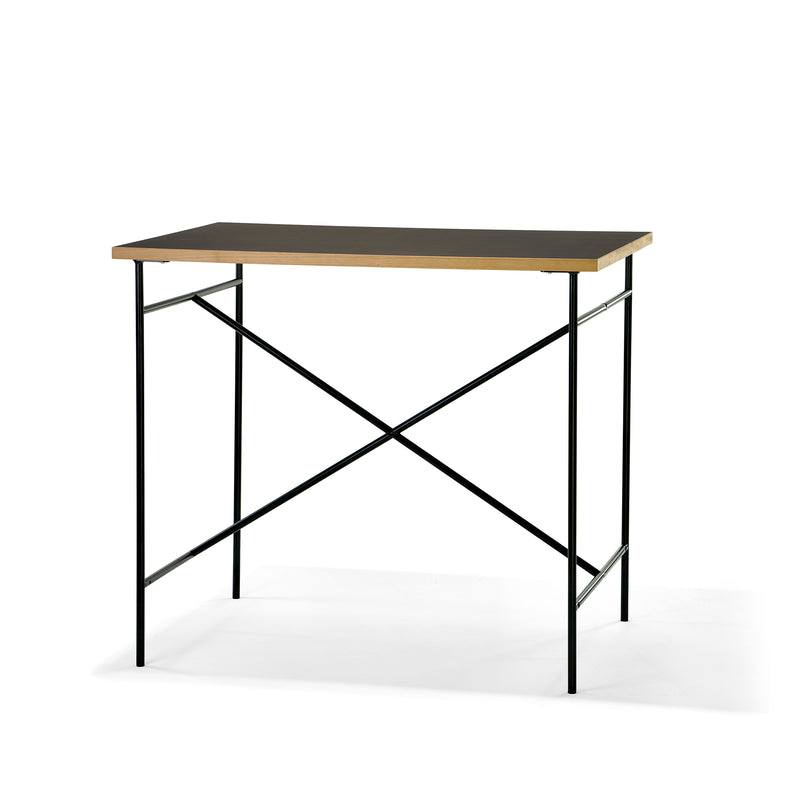 The Milla High Table 1100 by Richard Lampert is a minimalist desk with a thin wooden tabletop and black metal frame, featuring cross-bracing for stability. Its sleek, modern design makes it ideal for small workspaces or home offices.