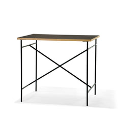 The Milla High Table 1100 by Richard Lampert is a minimalist desk with a thin wooden tabletop and black metal frame, featuring cross-bracing for stability. Its sleek, modern design makes it ideal for small workspaces or home offices.