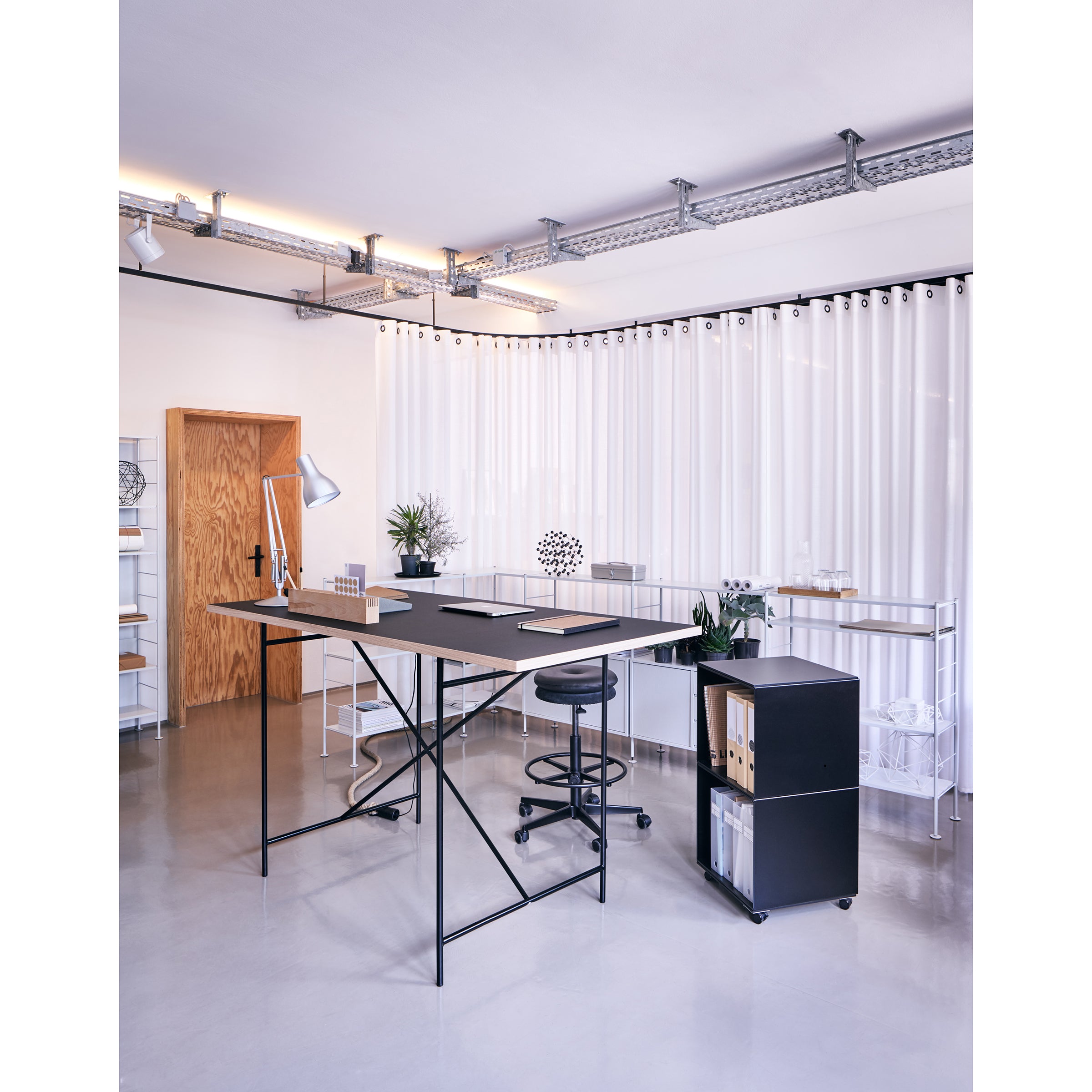 A modern, minimalistic office featuring a Richard Lampert Milla High Table 1100, stool, and rolling cart. The room includes a wooden door, white shelves, plants, artwork, and ceiling lights. The bright space has a glossy floor and pleated white curtains.