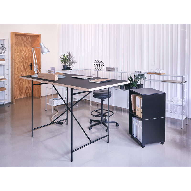 The office features a large, modern desk with the sleek Mr. Round High Chair by Richard Lampert. Nearby, a black filing cabinet and shelves store office supplies. The room is adorned with white curtains, a wooden door, and plants for decoration.