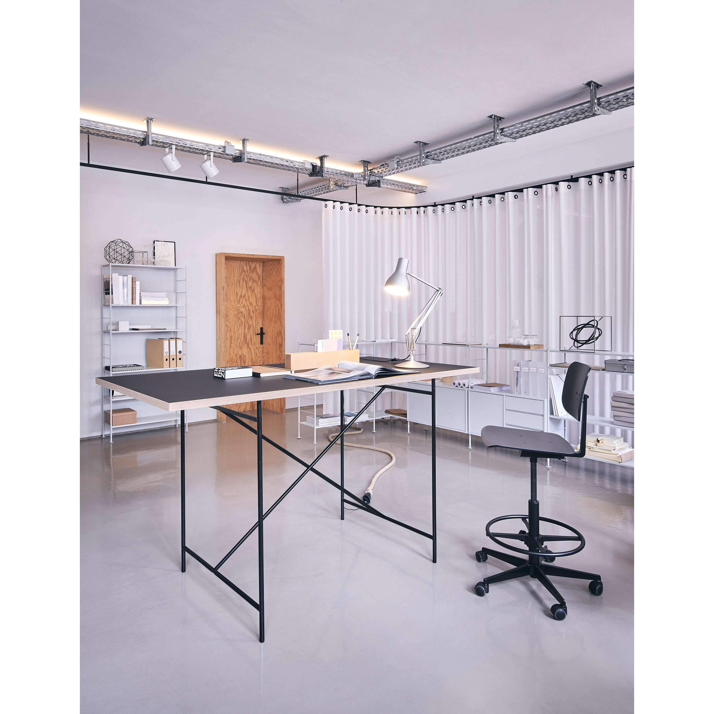 A modern office features a sleek black table, the Mr. Square High Chair by Richard Lampert for superior comfort, a desk lamp, and various supplies. The room includes white walls, vertical blinds, and a wooden door with shelves of books and decor in the background.