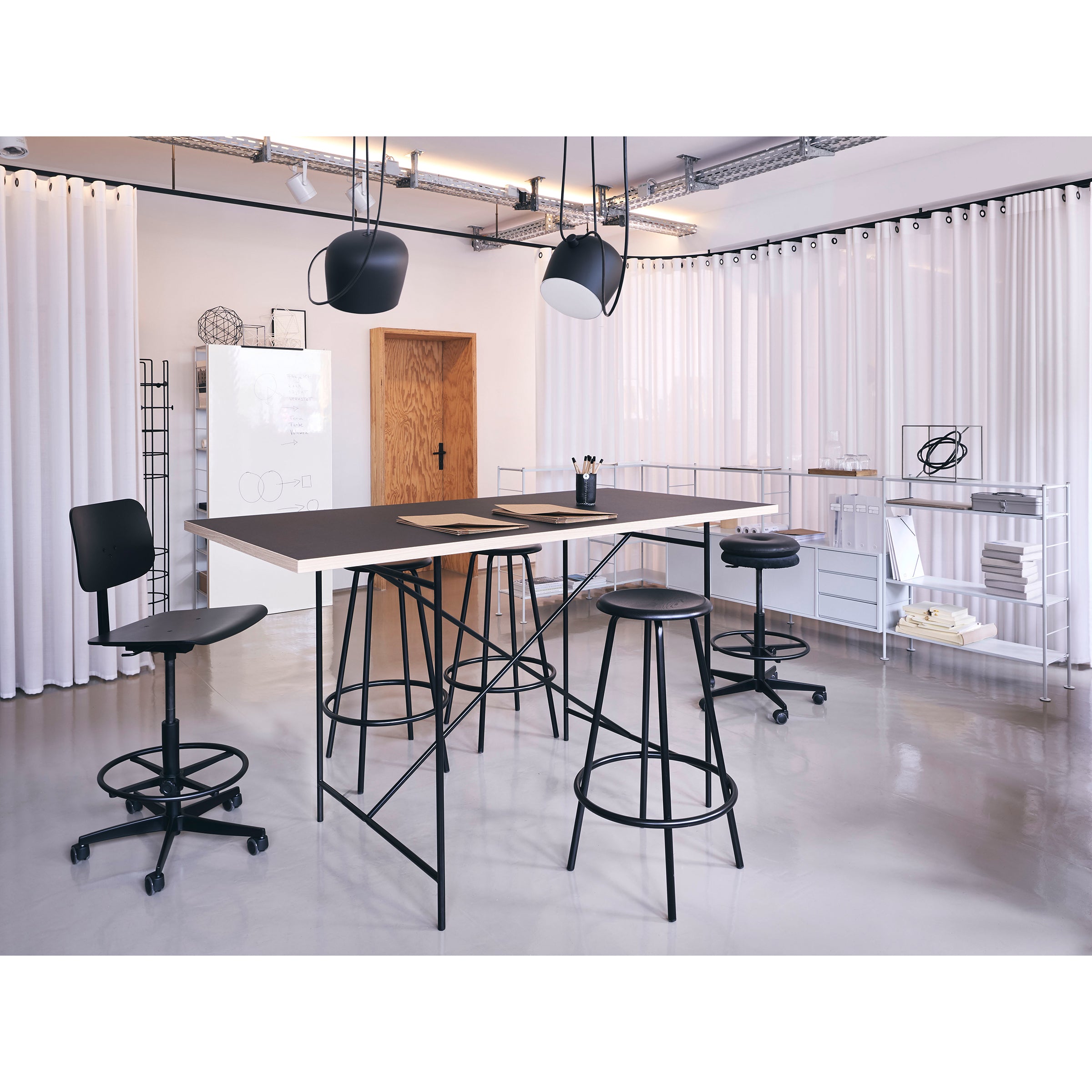 A modern office showcases a sleek black table paired with Mr. Round High Chair by Richard Lampert, featuring space-saving adjustable stools and a swivel chair. Minimalist decor includes white curtains, shelves, and pendant lights, complemented by a corkboard and supplies in the background.