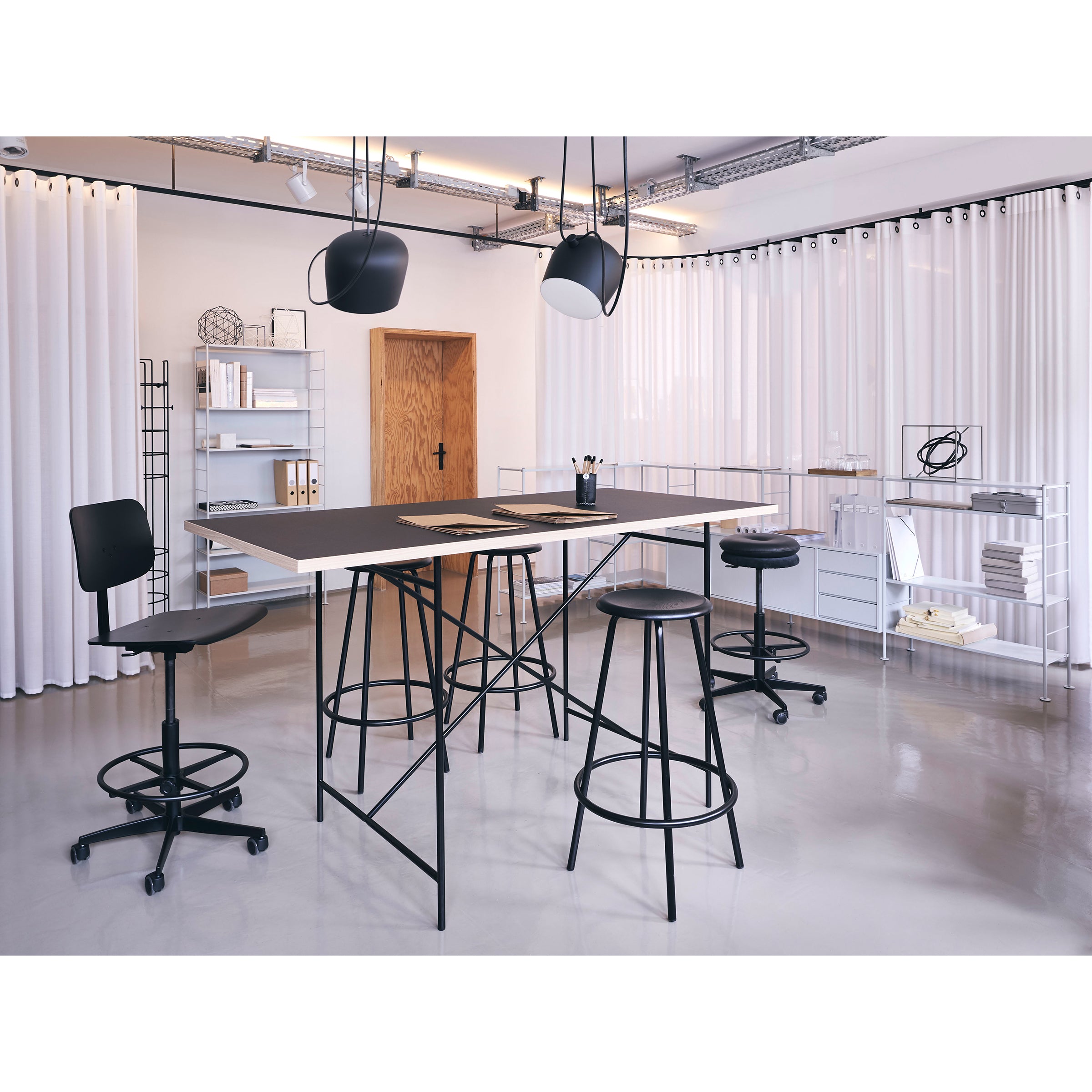A modern office features high desks and bar stools, two black chairs, white shelves, geometric lights, and white curtains. The Richard Lampert Milla High Table 1100 adds style to the minimalist setup, complemented by a wooden door and various office supplies.