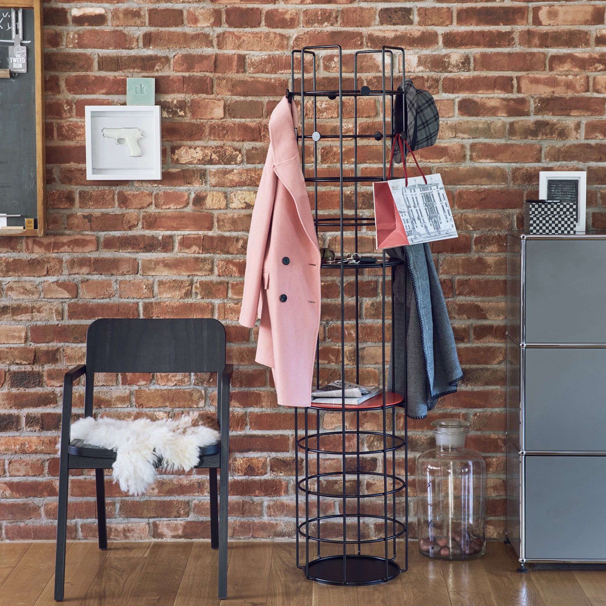 The setup features Richard Lamperts Bazar Coat Stand, a stylish black wireframe unit holding a pink coat, scarf, hat, and paper bag against a brick wall backdrop. To the left is a black chair accented with a white fur throw under small framed artwork, blending fashion with practical storage.