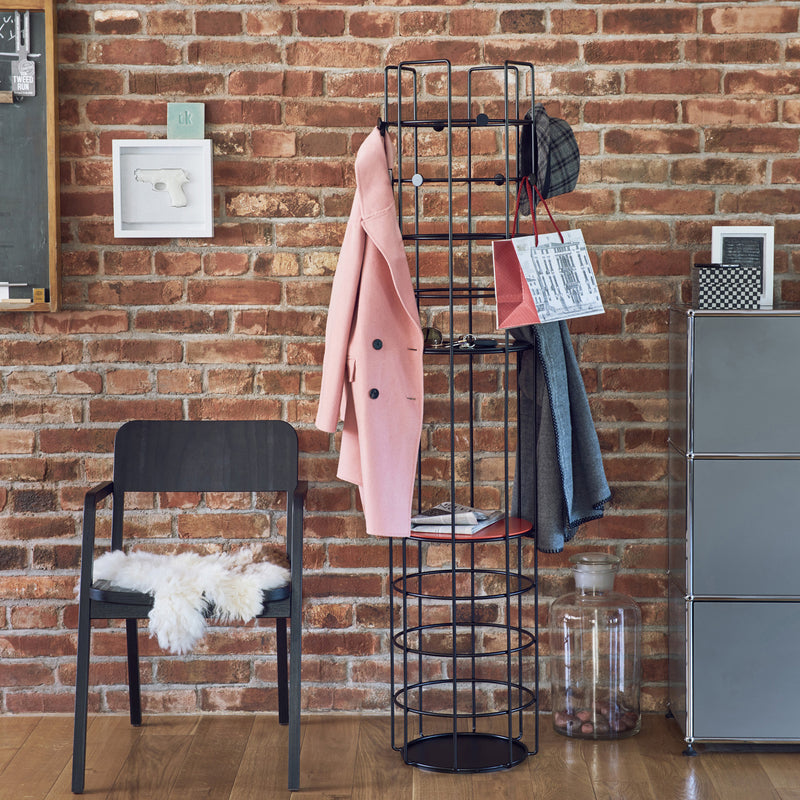 The setup features Richard Lamperts Bazar Coat Stand, a stylish black wireframe unit holding a pink coat, scarf, hat, and paper bag against a brick wall backdrop. To the left is a black chair accented with a white fur throw under small framed artwork, blending fashion with practical storage.