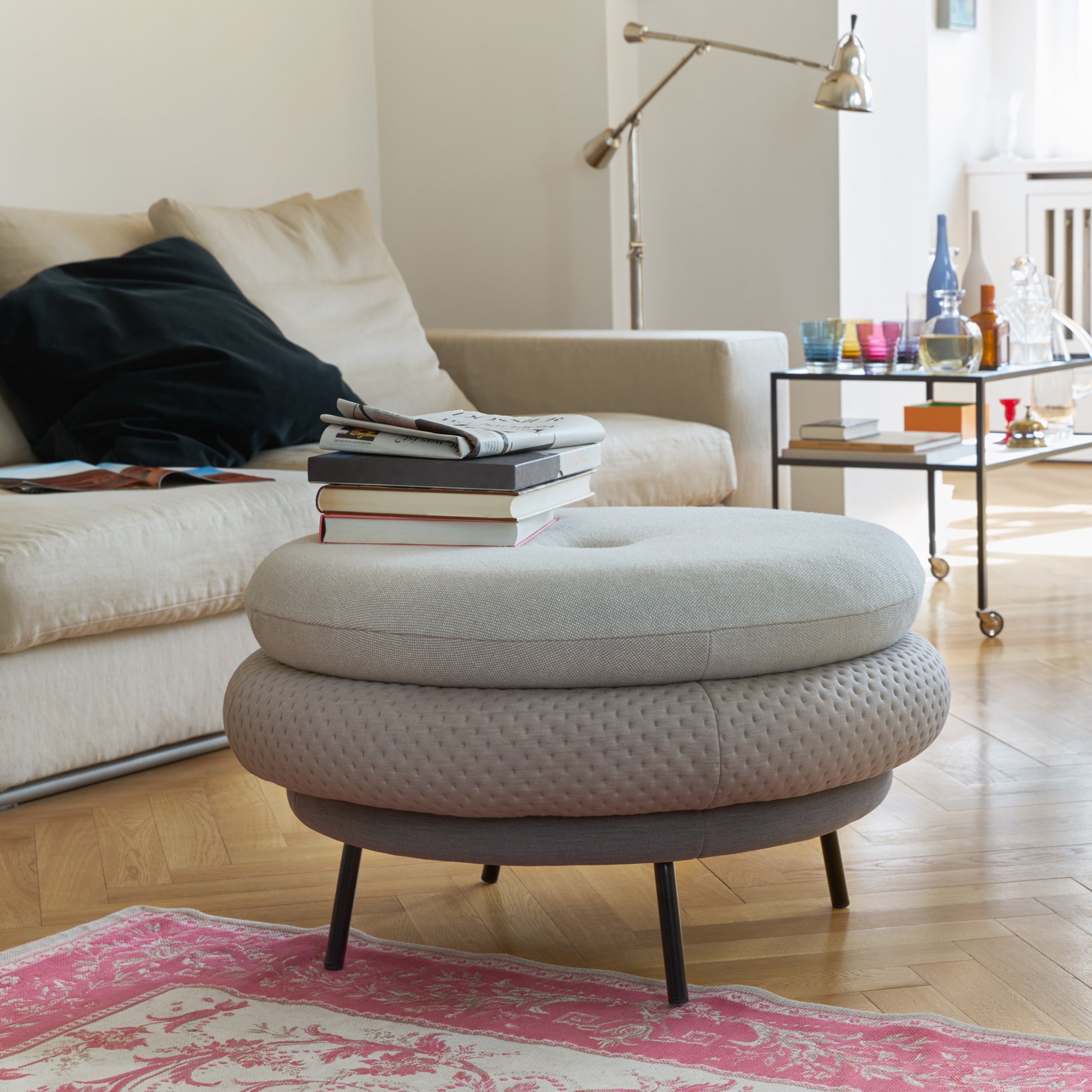 A cozy living room features a light beige sofa with combinable covers, a stack of books on a round ottoman, and a side table holding glasses and decor. Showcasing Fat Tom by Richard Lampert, the space includes wooden flooring, a pink patterned rug, and a standing lamp in the background.