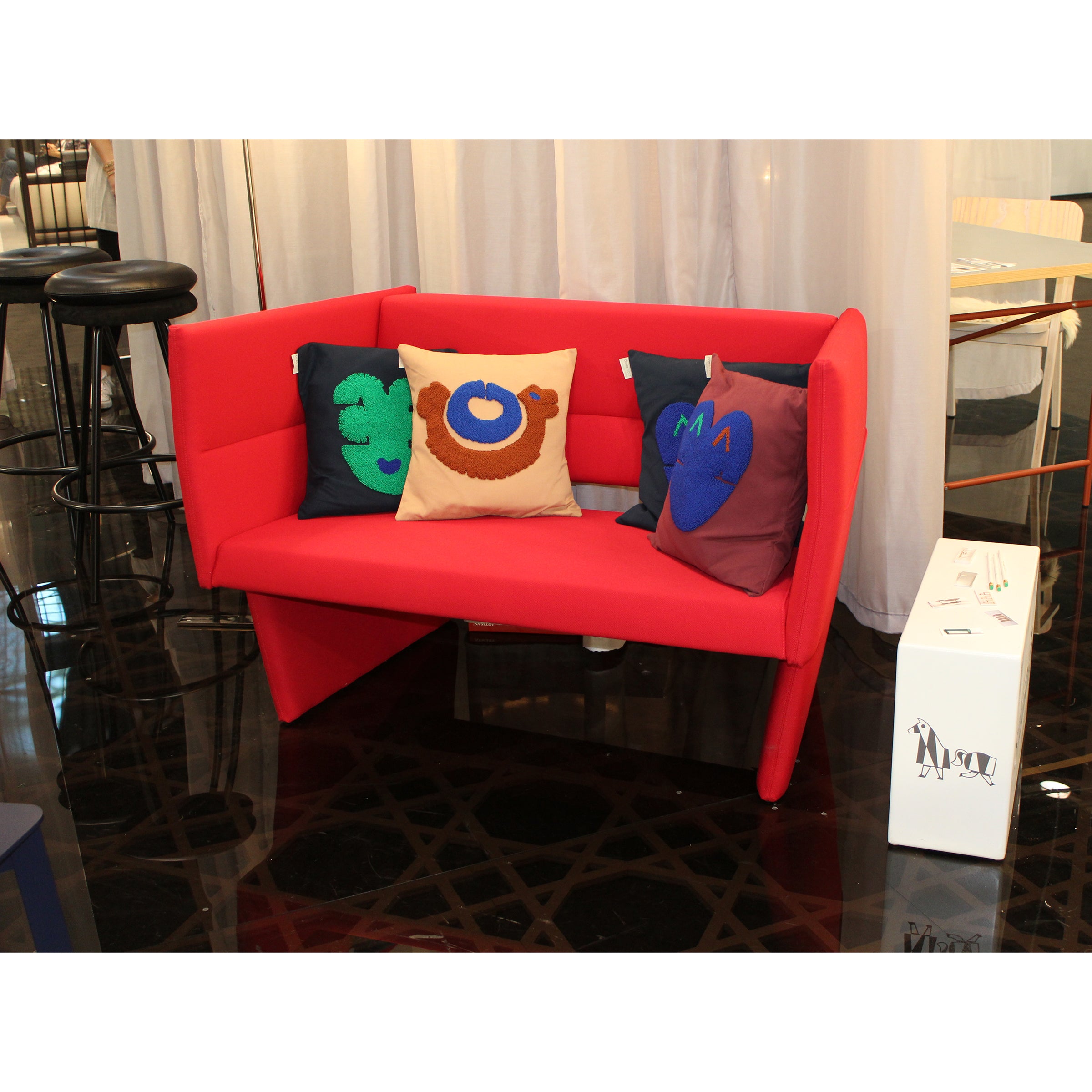 The Cup Sofa by Richard Lampert, sporting a modern red hue and triangular base, is accented with assorted colorful cushions. It rests on a glossy black floor amid white curtains and nearby furniture such as a black bar stool and a white box.