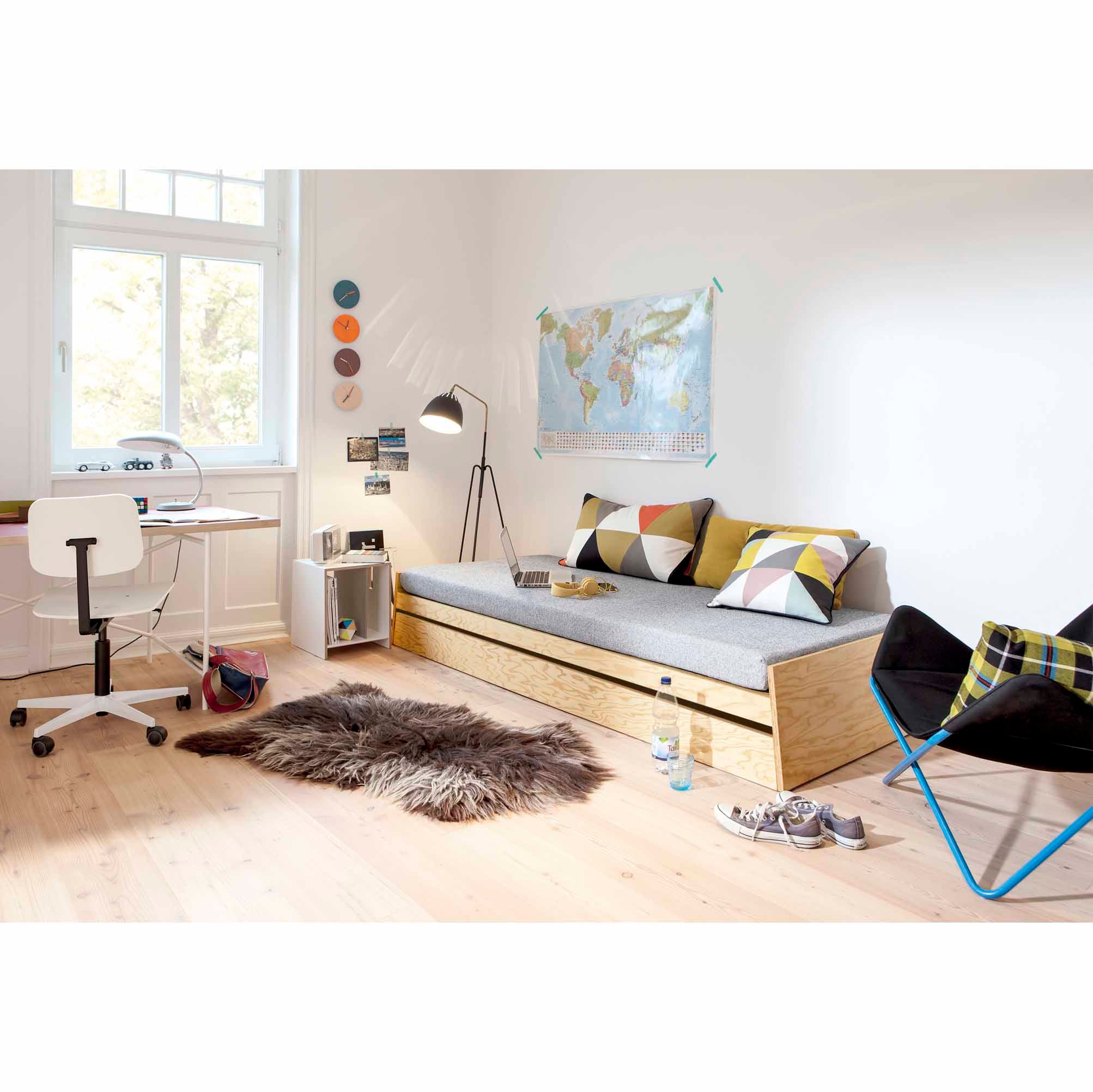 A bright room by Jochen Gros includes a wooden daybed with geometric cushions, a world map, a desk with a white chair, and an aluminum tripod floor lamp. An Alu Alu Wallclock by Richard Lampert sits on the small shelf as natural light floods the fuzzy rug and blue chair.