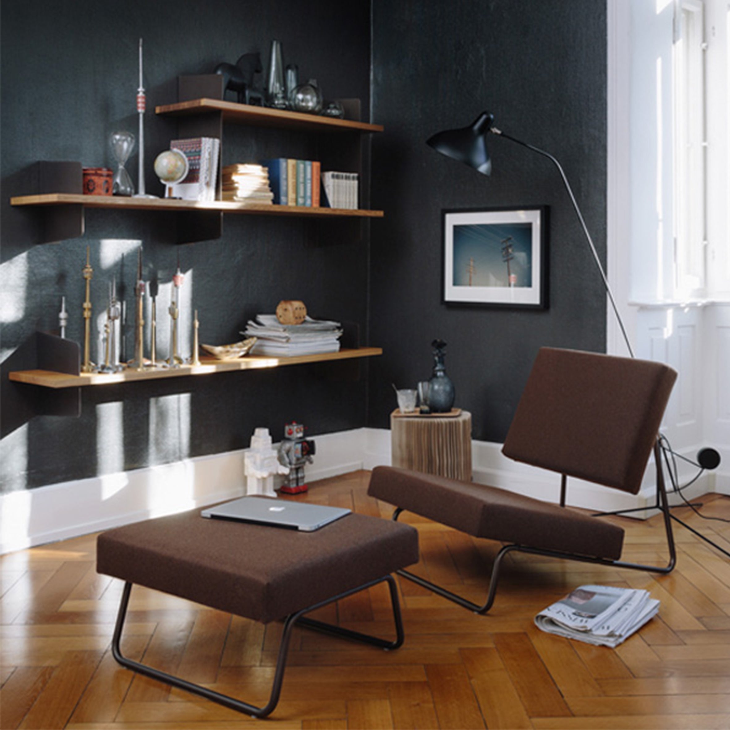 A modern living room with dark walls and wooden floors showcases sophisticated furniture, featuring the Atelier chair and ottoman by Richard Lampert, a large black floor lamp, and wooden shelves adorned with books and decor. Sunlight floods through the window, enhancing the cozy atmosphere.