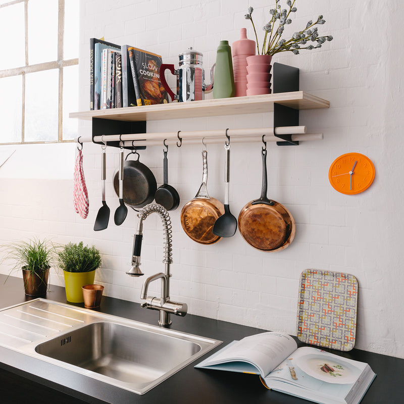 A modern kitchen setup with Richard Lamperts Atelier stainless steel sink and faucet. Above, wooden shelves with steel brackets display books, vases, and plants. Kitchen utensils and pans hang below. A bright orange wall clock complements a colorful cutting board and open book on the counter.