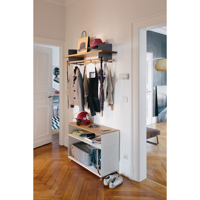 A tidy hallway is adorned with the Richard Lampert Atelier wall-mounted coat rack holding jackets, scarves, and storage boxes. Beneath it stands a white console with sophisticated design elements displaying a red helmet, baskets, and magazines. Shoes are neatly aligned on the wooden parquet floor in this well-lit, modern space.