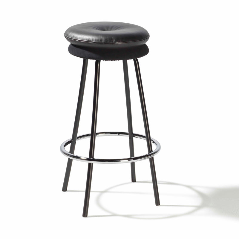 Introducing the Big Tom Barstool by Richard Lampert: a modern black stool with a round cushioned seat in leather and textile, supported by four tubular steel legs connected by a circular metal footrest, elegantly casting a shadow on a pristine white background.