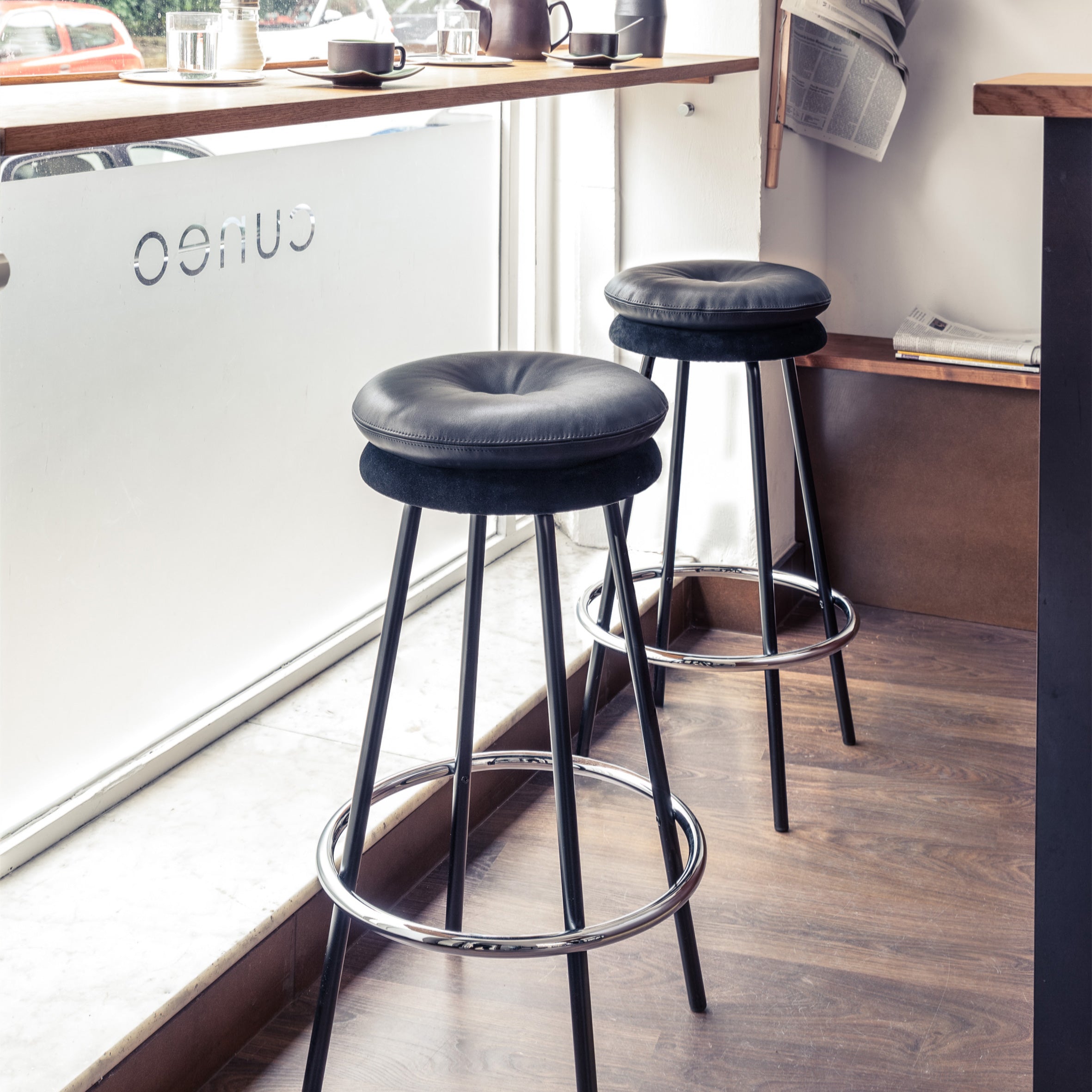 Two Richard Lampert Big Tom Barstools, featuring tubular steel frames and black leather covers, stand by a cafe window. Nearby, a wooden shelf holds newspapers and coffee cups. OENO is reverse-printed on the glass, while the wooden floor adds warmth to this cozy setting.