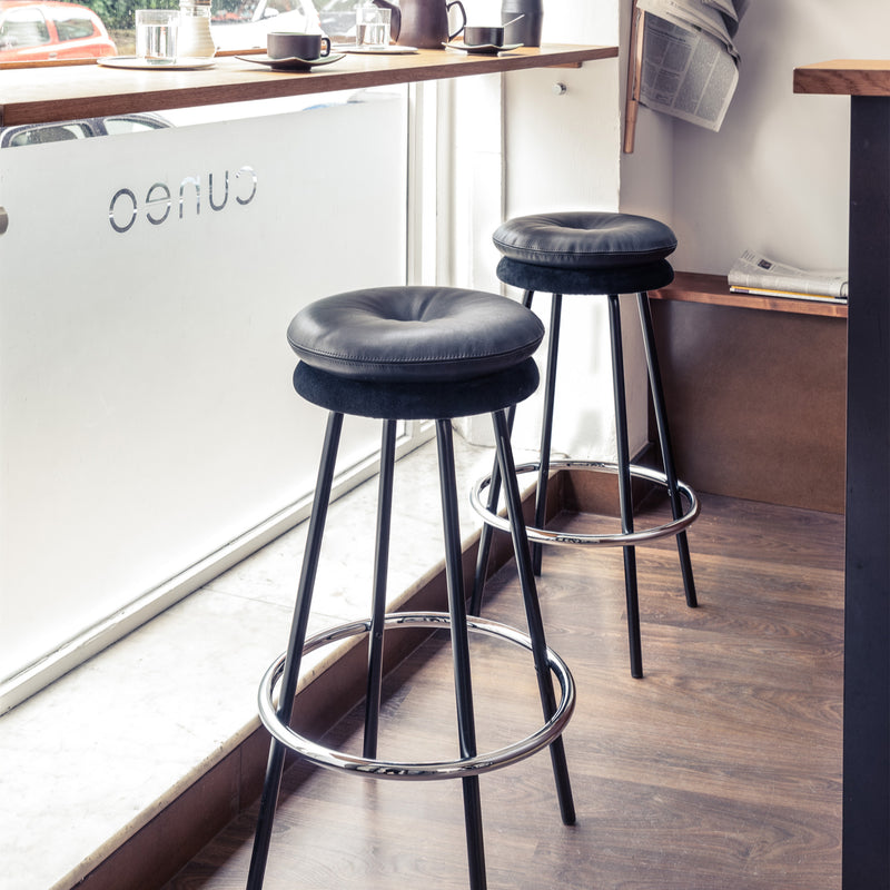 Two Richard Lampert Big Tom Barstools, featuring tubular steel frames and black leather covers, stand by a cafe window. Nearby, a wooden shelf holds newspapers and coffee cups. OENO is reverse-printed on the glass, while the wooden floor adds warmth to this cozy setting.