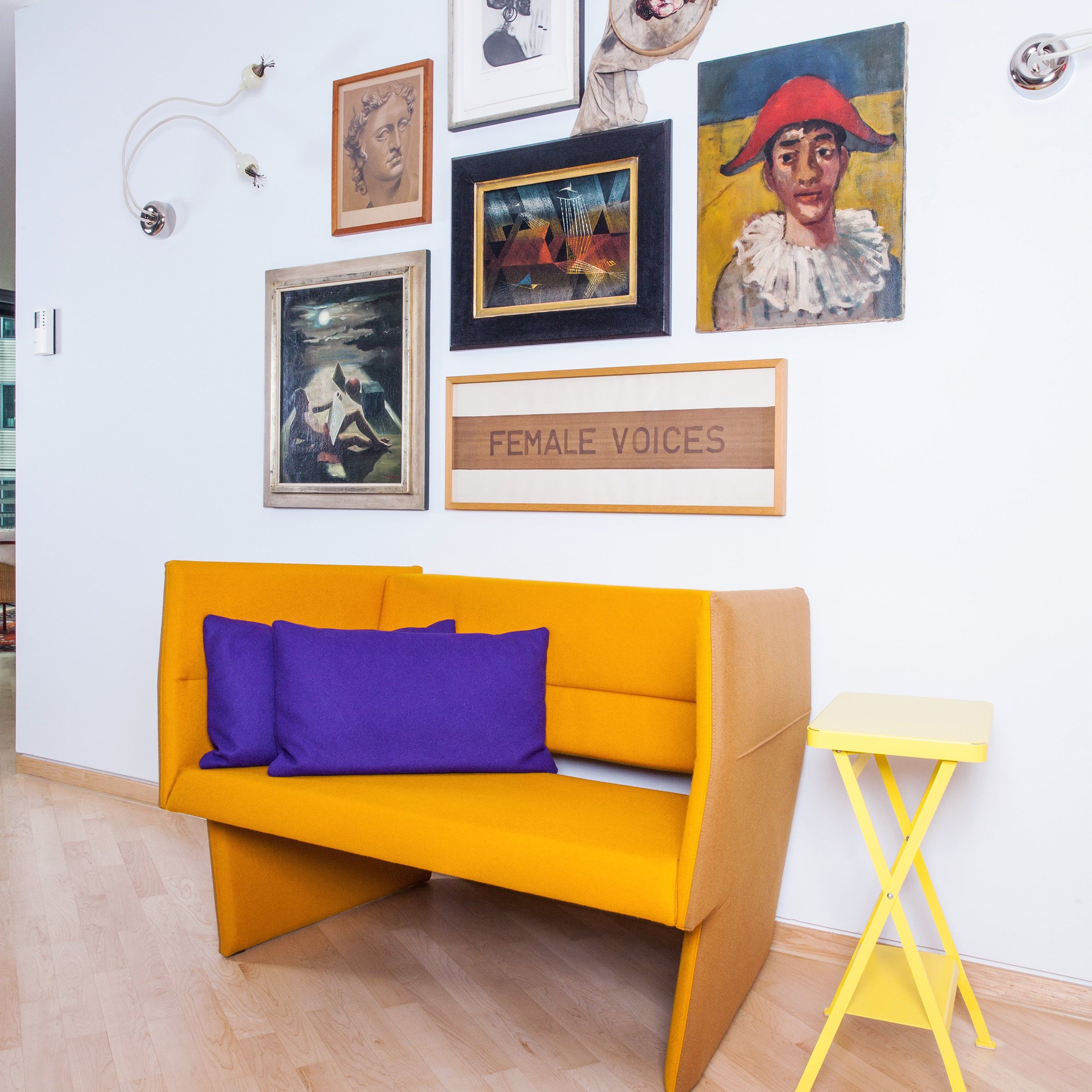 A chic room showcases the Richard Lampert Cup Sofa with high-end upholstery in mustard and purple cushions, next to a bright yellow side table. The white wall above is decorated with diverse artwork and a FEMALE VOICES sign, while the light wooden floor enhances this avant-garde design space.