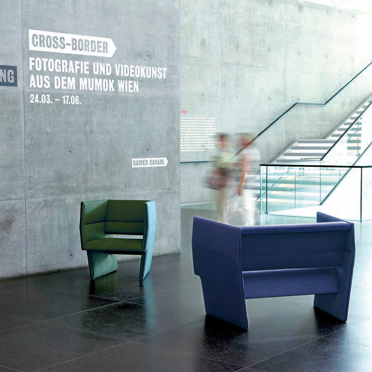 A sleek, dark floor hosts two angular Cup Sofas by Richard Lampert in blue and green high-end upholstery. A blurred couple strolls past a concrete wall with German text about a Mumok Wien exhibition, while a staircase with a glass railing is visible in the background.