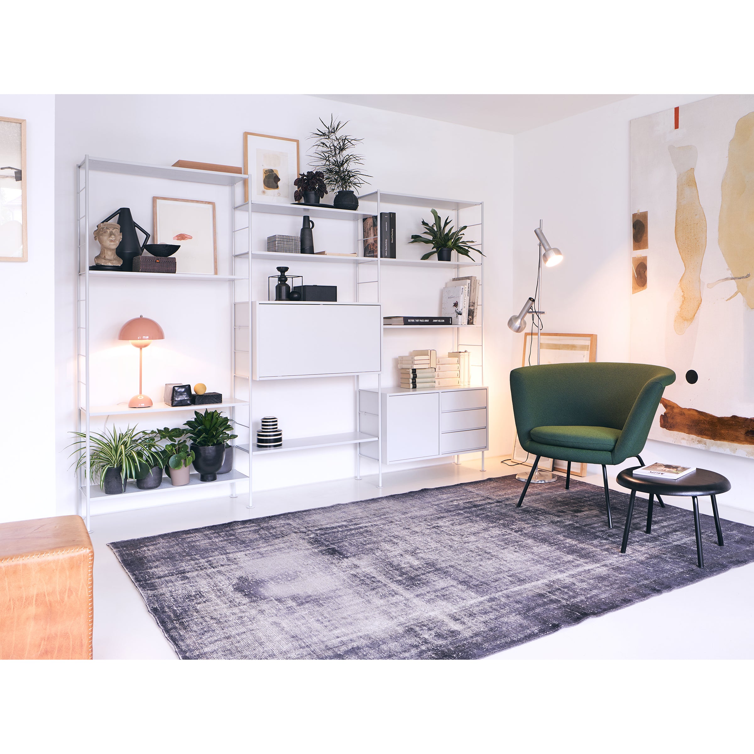 A modern living room displays mid-century design with white shelving filled with books, plants, and decor. The Richard Lampert H 57 Lounge Chair and matching footstool sit on a gray rug near a tall floor lamp. Abstract art adorns the white walls, enhancing the timeless appeal.