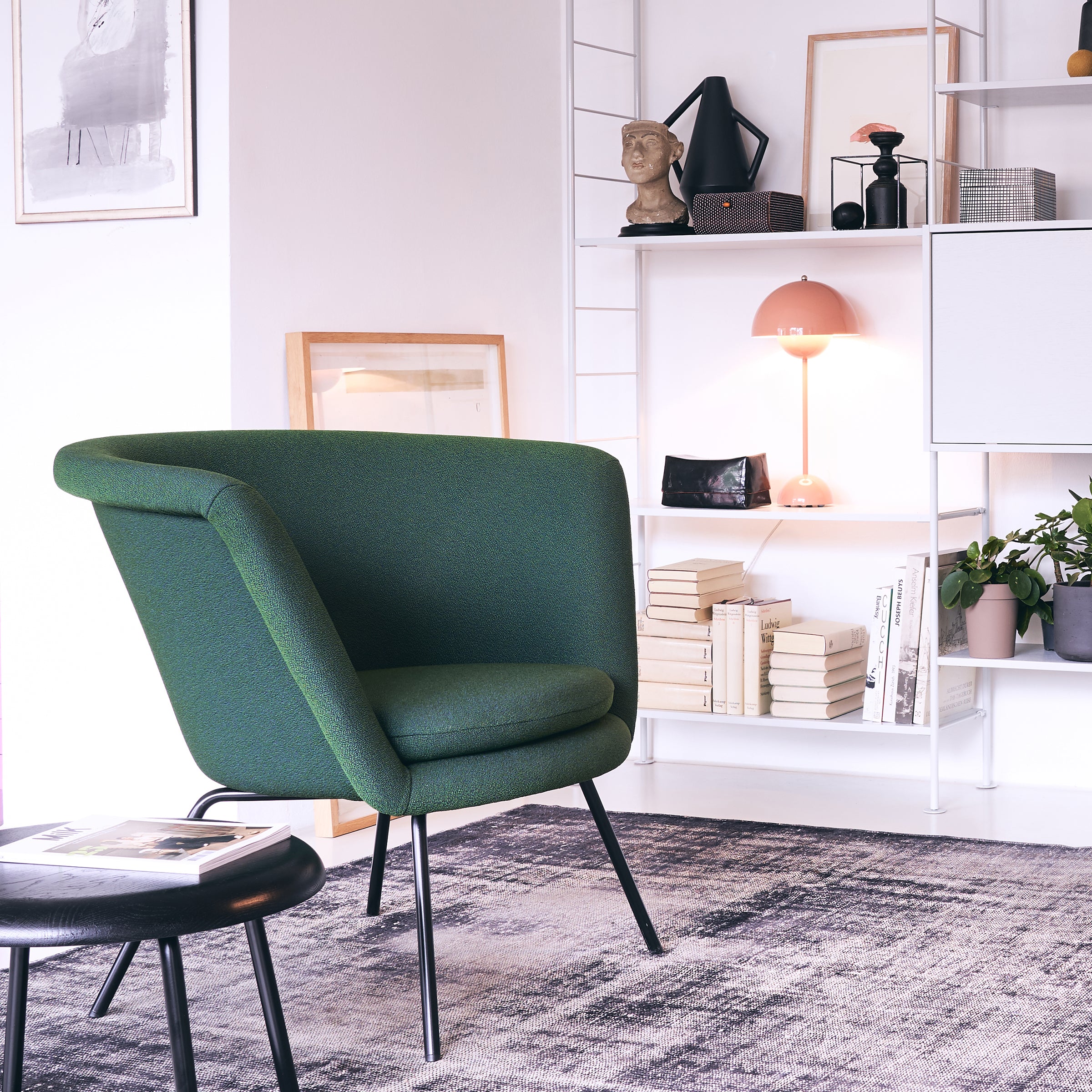 A modern living room showcases mid-century design with a green upholstered H 57 Lounge Chair by Richard Lampert, a black round side table, and an abstract-patterned rug. The white shelving unit displays books and art pieces while a pink lamp adds color, inspired by Herbert Hirche furniture.