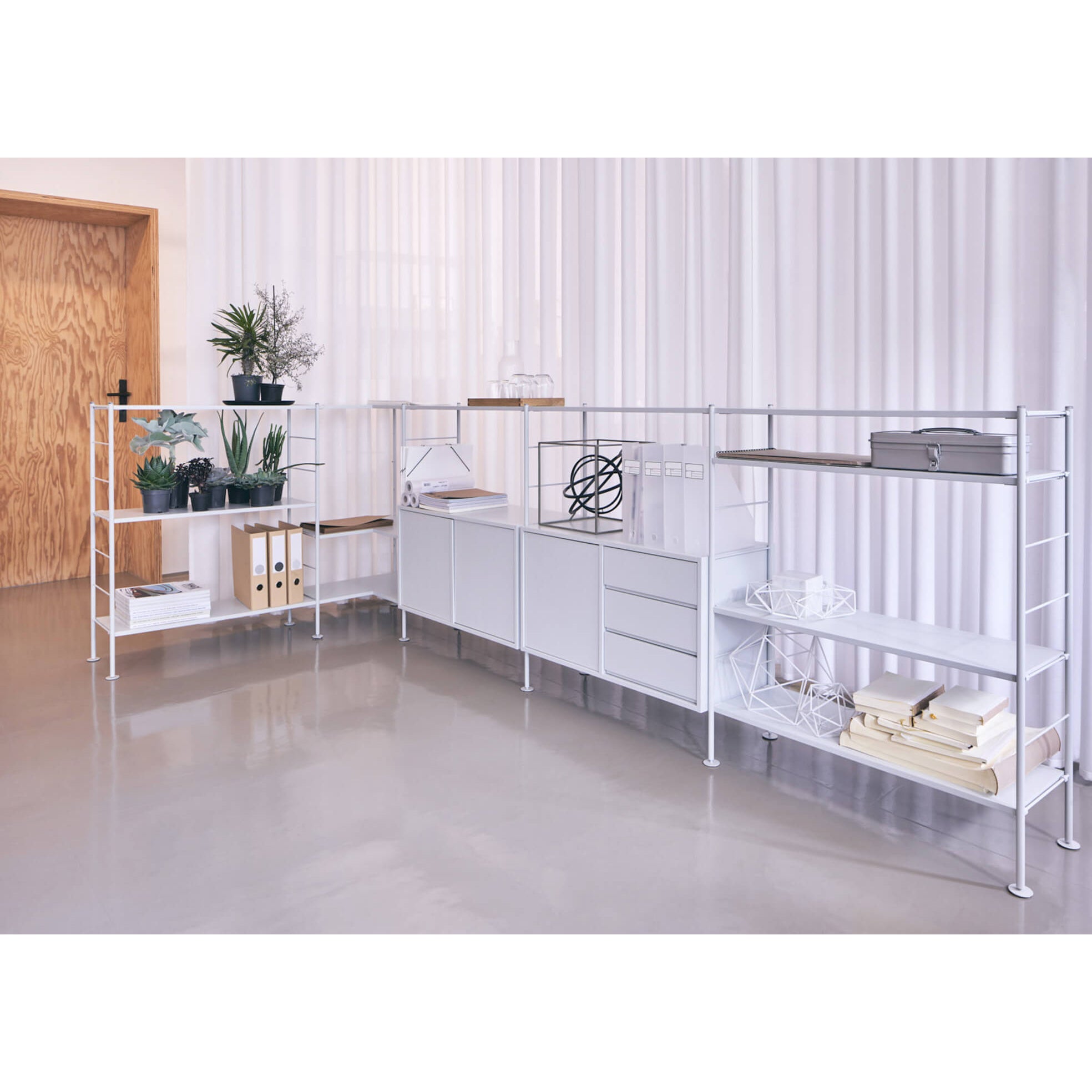 The DHS10 Shelf by Richard Lampert showcases German industrial design in a modern room, displaying potted plants, books, and decor. The polished concrete floor complements white curtains and a wooden door in this sleek shelving system.
