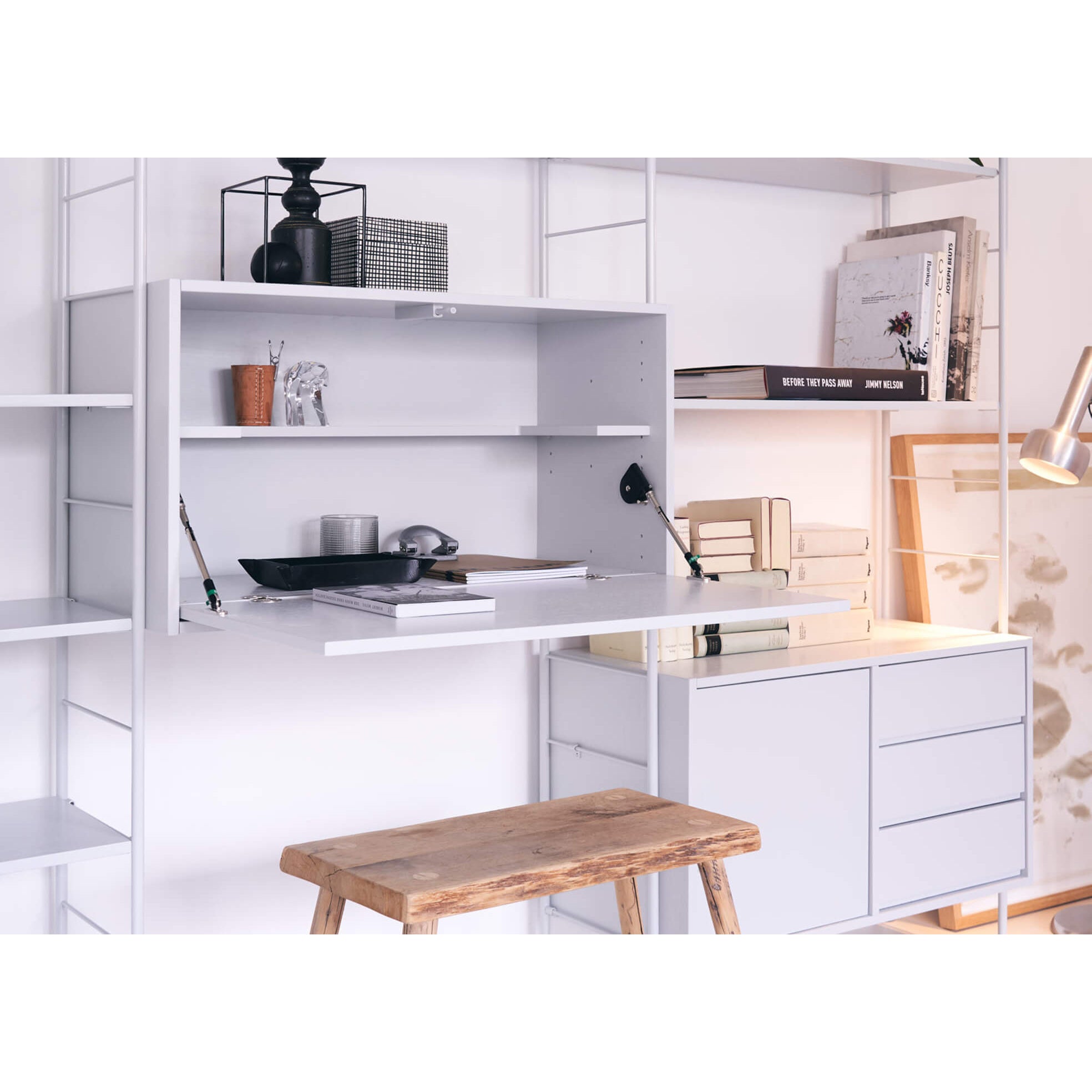 The DHS10 Shelf by Richard Lampert, a modern white shelving system showcasing German industrial design, features books and decor like a black vase and metal bowl. A wooden stool complements the fold-down desk section, while a lamp adds warm light to enhance the cozy atmosphere.