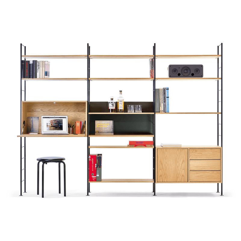 The DHS10 Shelf by Richard Lampert, a modular system in German industrial design, features wooden shelves with black supports. It displays books, a sound system, a laptop, and a liquor set alongside closed cabinets. A stool is placed in front of its dropdown desk section.