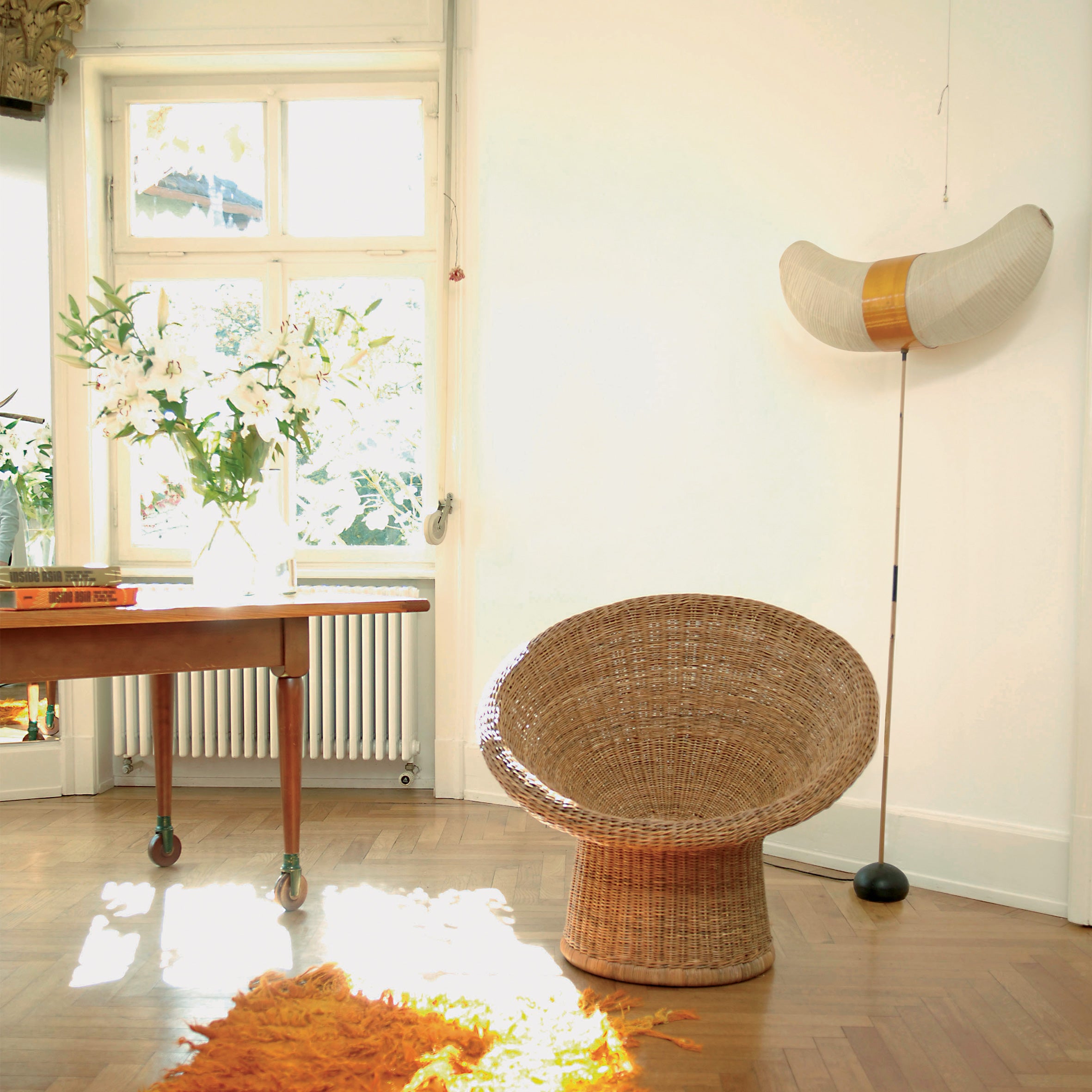 A sunlit room showcases a Richard Lampert E10 Lounge Chair in rattan, paired with an orange shaggy rug. A wooden table holds books and white flowers elegantly. A large, curved paper lamp by Egon Eiermann stands in the corner, while white walls and a window fill the space with natural light.