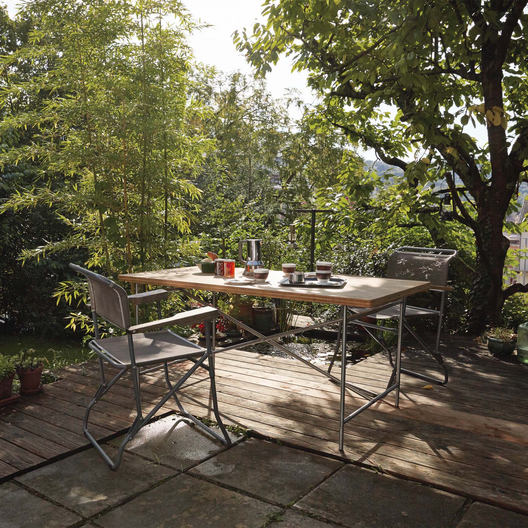A wooden deck showcases the Eiermann 2 Dining Table by Richard Lampert and two chairs set for breakfast amidst lush greenery. Sunlight filters through leaves, casting dappled shadows. A coffee pot and cups rest on the table—a serene scene echoing Egon Eiermann’s design ethos.