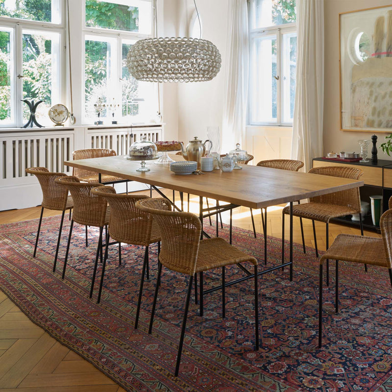 The dining room showcases the Richard Lampert Eiermann 2 Dining Table with ergonomic design, surrounded by wicker chairs on a patterned rug. A decorative chandelier hangs above, large windows with sheer curtains bring in light, and in the background, a sideboard and artwork complement the space.
