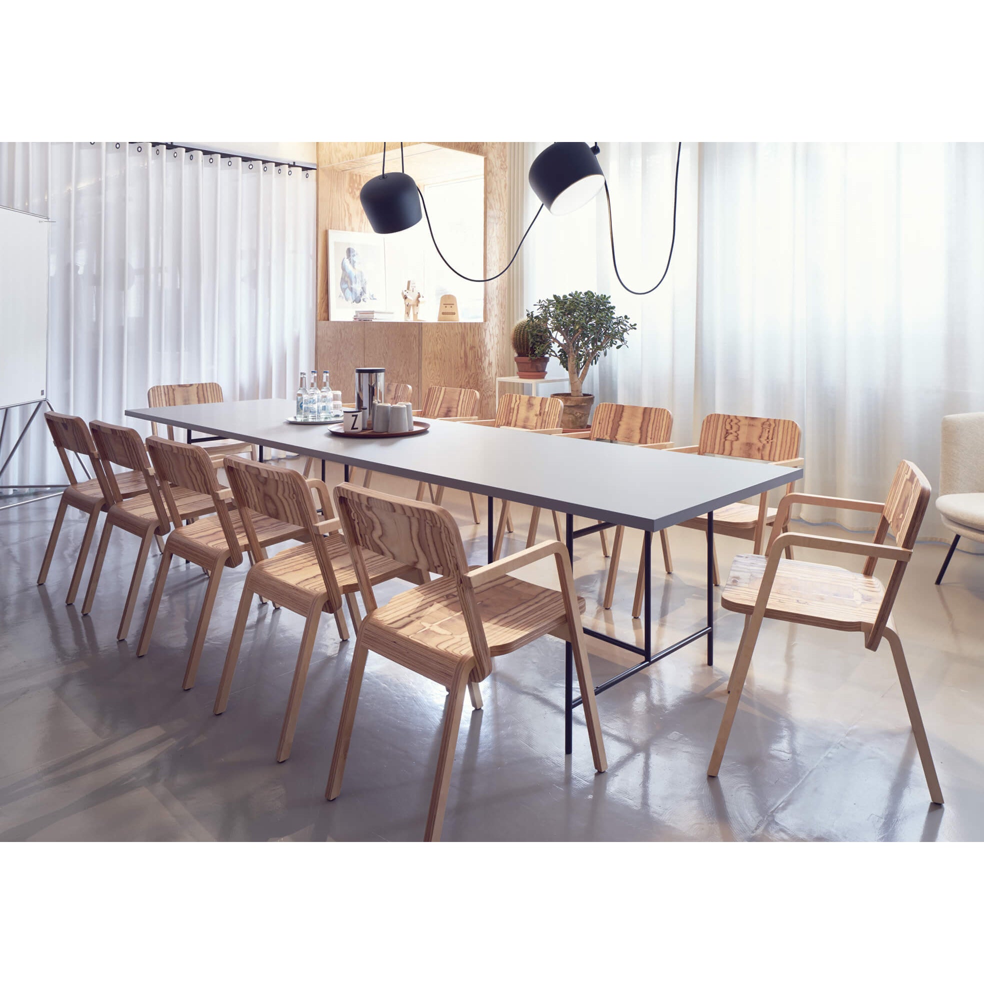 A modern conference room with a sleek Eiermann 3 Table by Richard Lampert, surrounded by wooden chairs. Two black pendant lights hang above, while a potted plant and artwork add flair to the white curtains and light-colored wall.