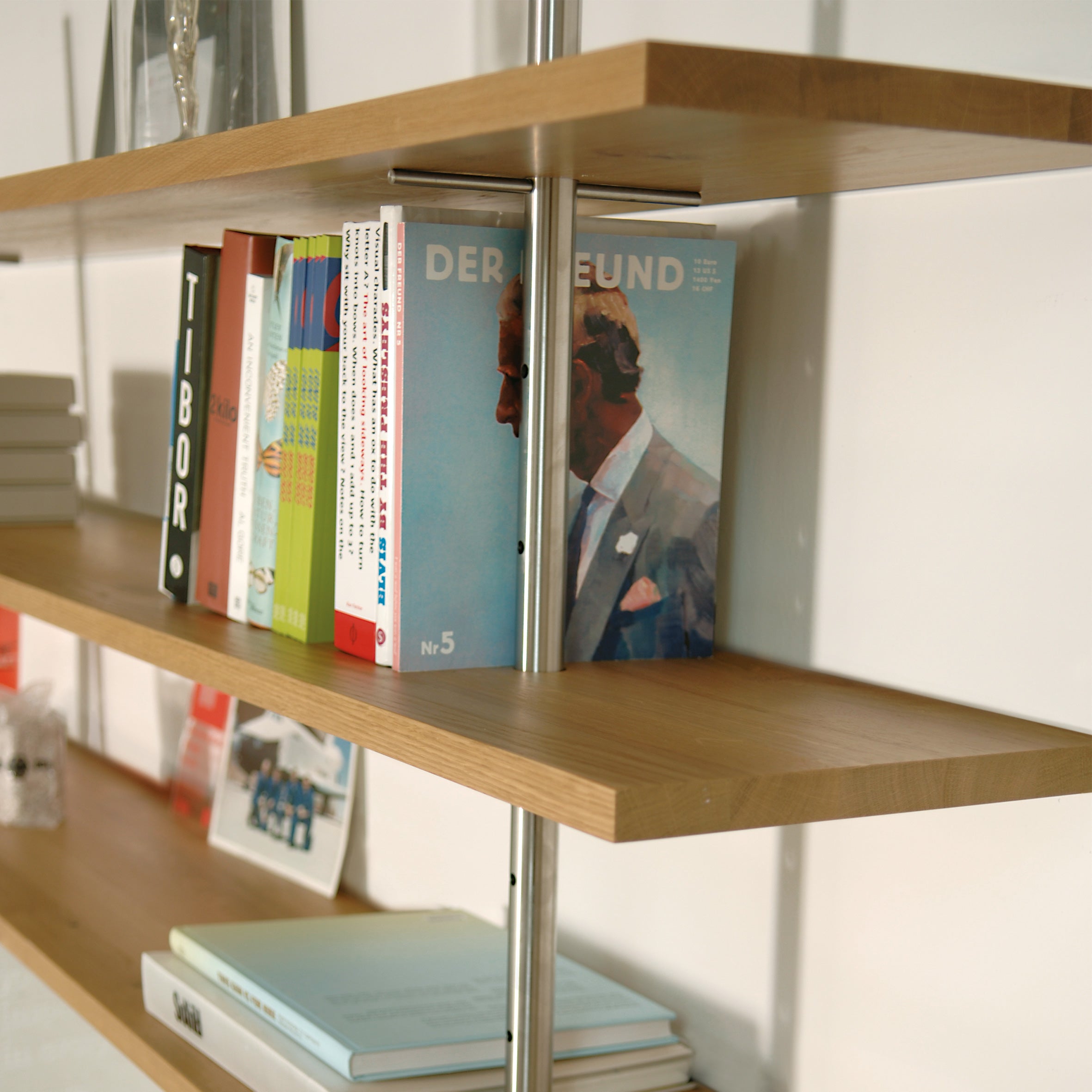 The Richard Lampert Eiermann Shelf, with wooden shelves and a classic metal frame, displays books and magazines vertically and horizontally, including one with an illustrated person in a suit.