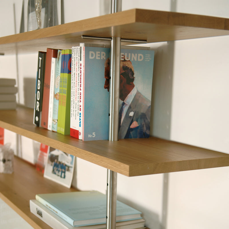 The Richard Lampert Eiermann Shelf, with wooden shelves and a classic metal frame, displays books and magazines vertically and horizontally, including one with an illustrated person in a suit.