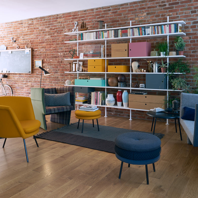 A cozy living room boasts an Egon Eiermann-inspired brick wall, a Richard Lampert Eiermann Shelf filled with books and decor, mustard and blue chairs, and a patterned rug on light wooden flooring. Partially visible on the left are a chalkboard and bicycle.