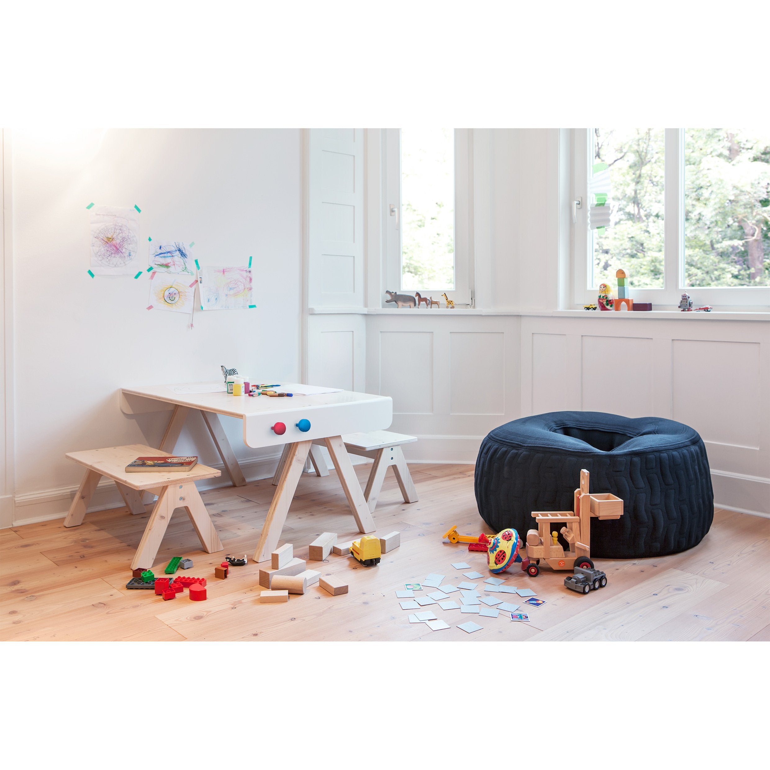 A bright playroom includes a white table, benches, scattered toys, wooden blocks, and a black bean bag chair. Richard Lamperts Famille Garage storage neatly organizes essentials. Childrens drawings adorn the walls while large windows let natural light illuminate the wooden floor.