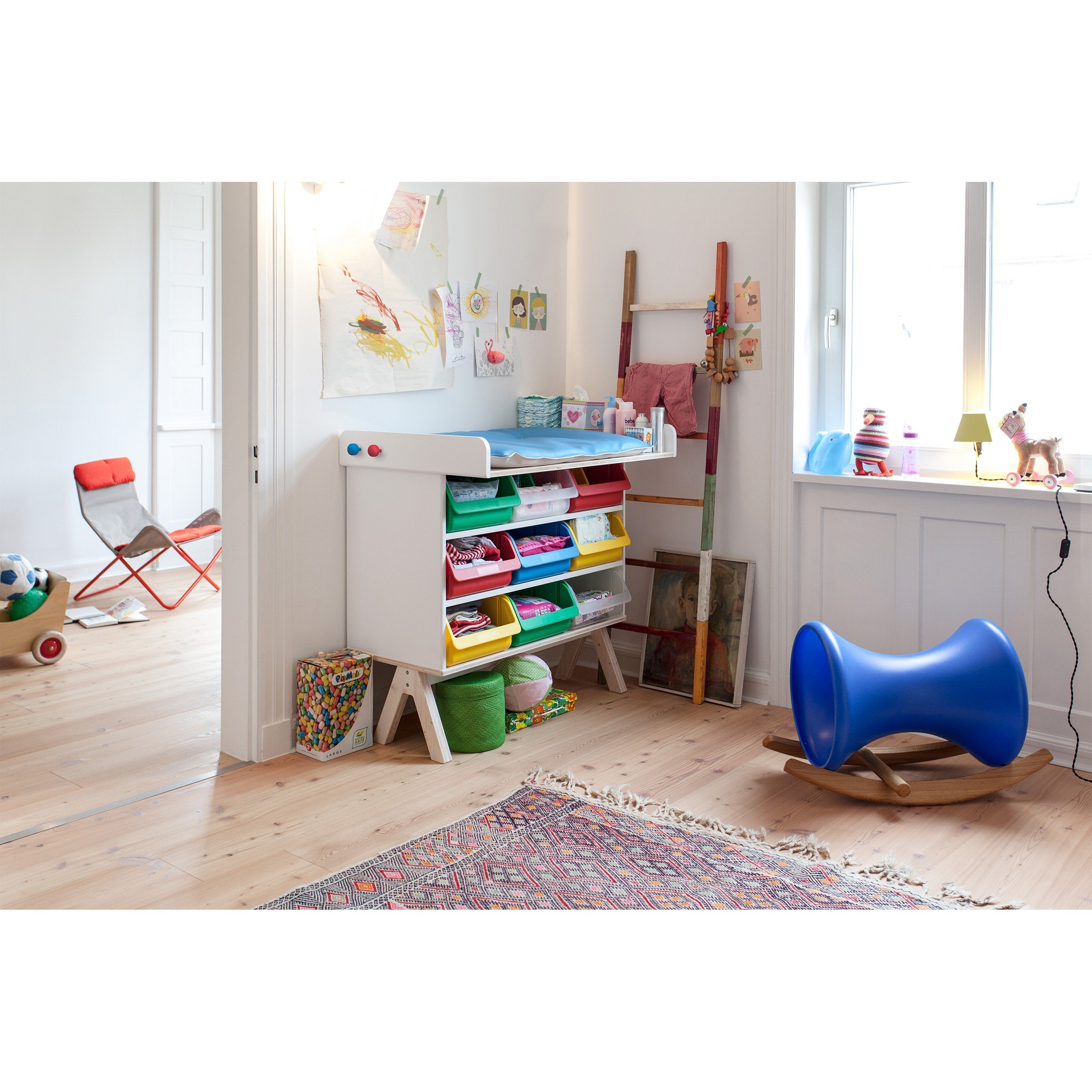 The childrens playroom features vibrant storage bins within the Richard Lampert Famille Garage unit, filled with toys. It includes a blue balance rocker, wall art, and a wooden ladder shelf. A red chair and basket sit in the adjacent room. Bright space with light wood flooring adds to its airy feel.