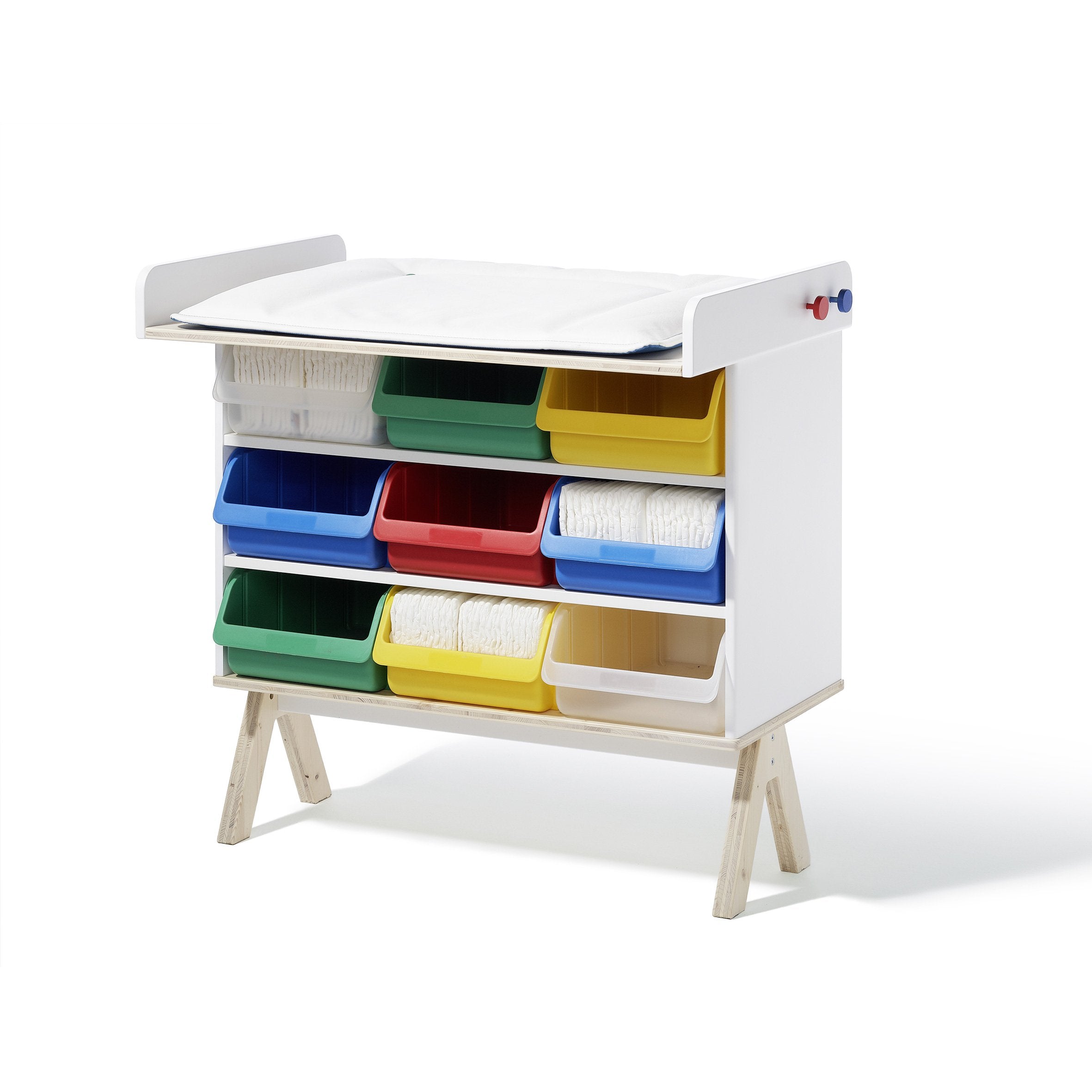 The Famille Garage - Storage by Richard Lampert is a childrens furniture system with a white frame, featuring colorful bins in blue, red, yellow, and green across multiple shelves. White paper rolls rest on top and inside some bins. It stands on wooden legs.