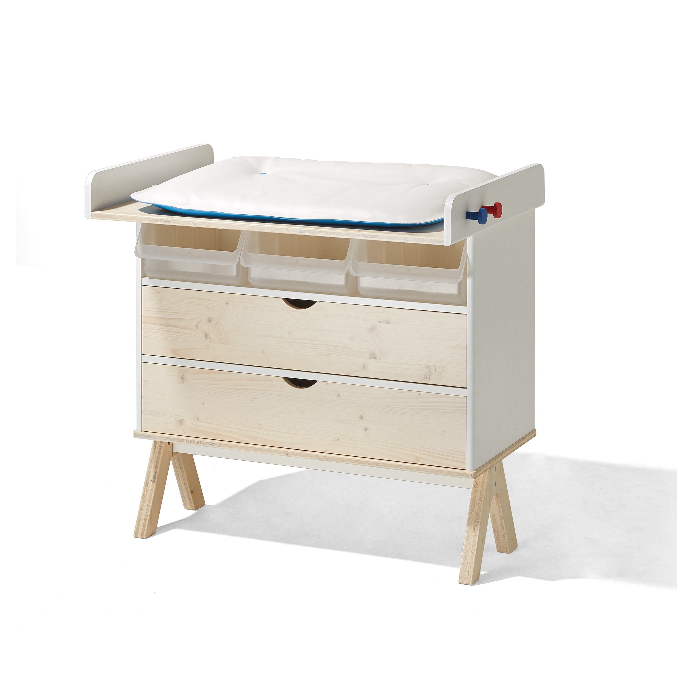 The Famille Garage by Richard Lampert is a versatile wooden changing table and storage unit, featuring light wood finish, two drawers, three open shelves with bins, a padded top changing mat, and red and blue knobs for a playful touch that complements any minimalist decor.