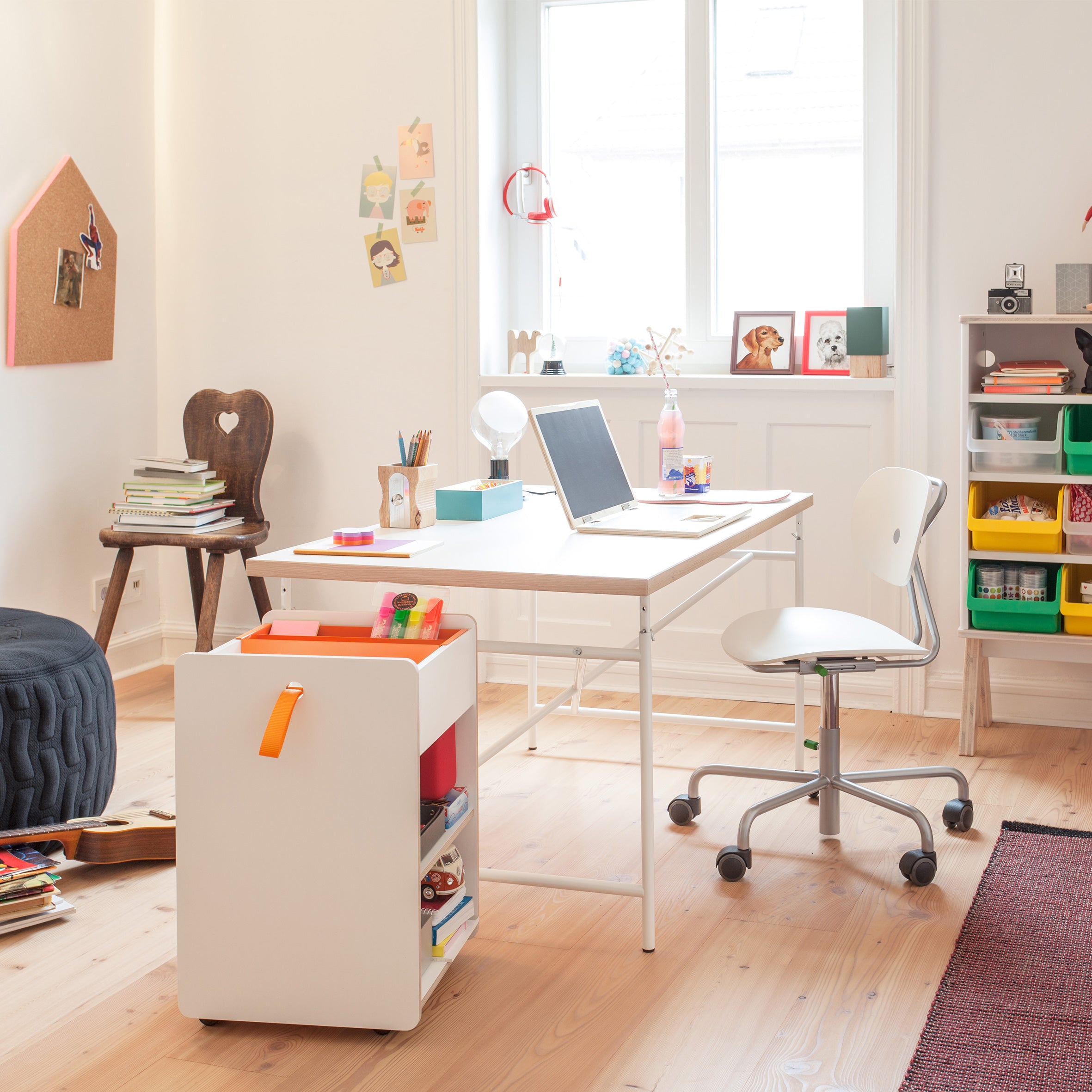 A bright study room features a desk with a laptop, books, and stationery near a window. A mobile cart and colorful storage bins are also present. Various items decorate the space, including Richard Lamperts Trolley Fixx Kids with secret compartments for your essentials.