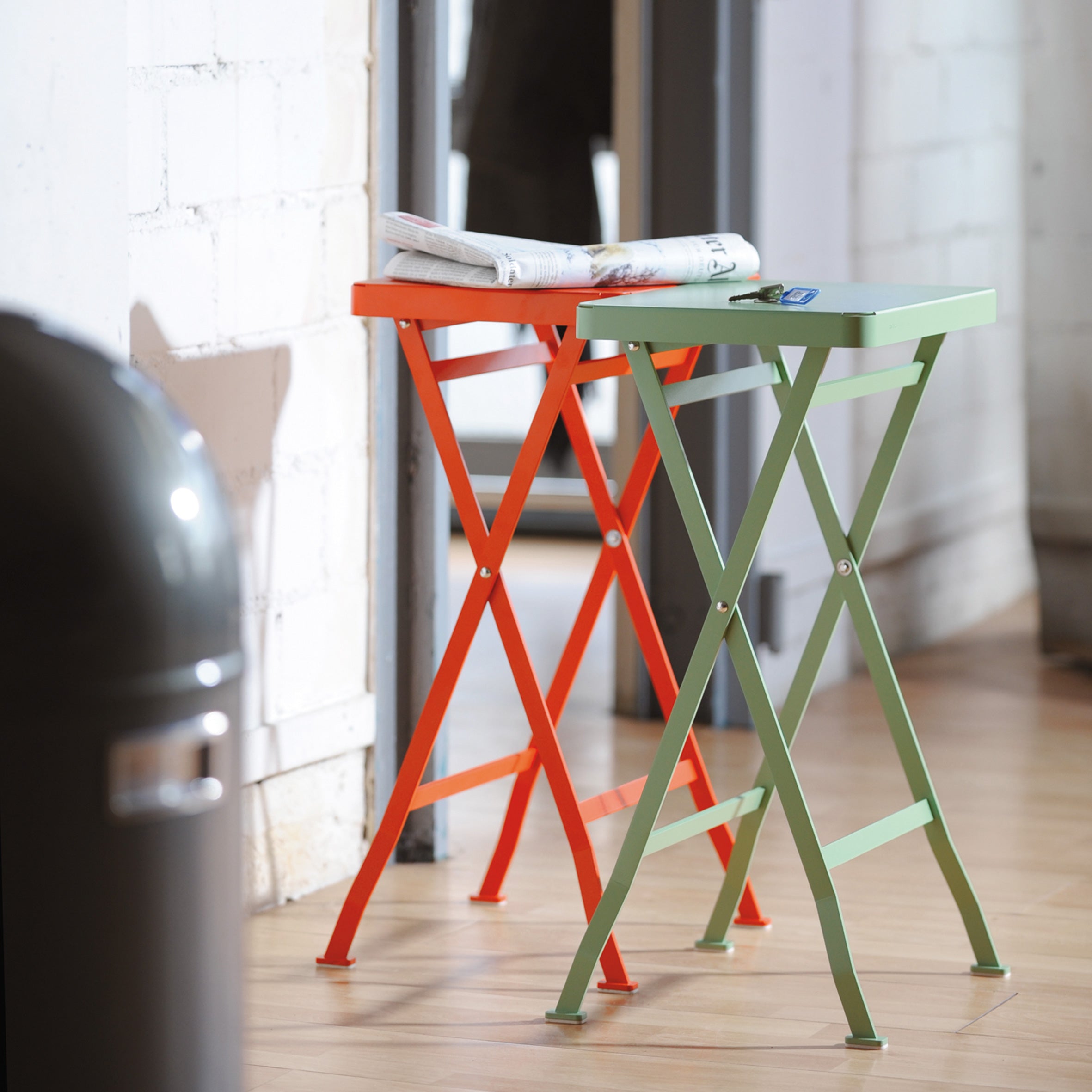 Two premium Flip Side Tables by Richard Lampert, one orange and one green, stand side by side against a white brick wall. A newspaper rests on the orange table. The setting is indoors with wooden flooring and a trash can partially visible in the foreground.