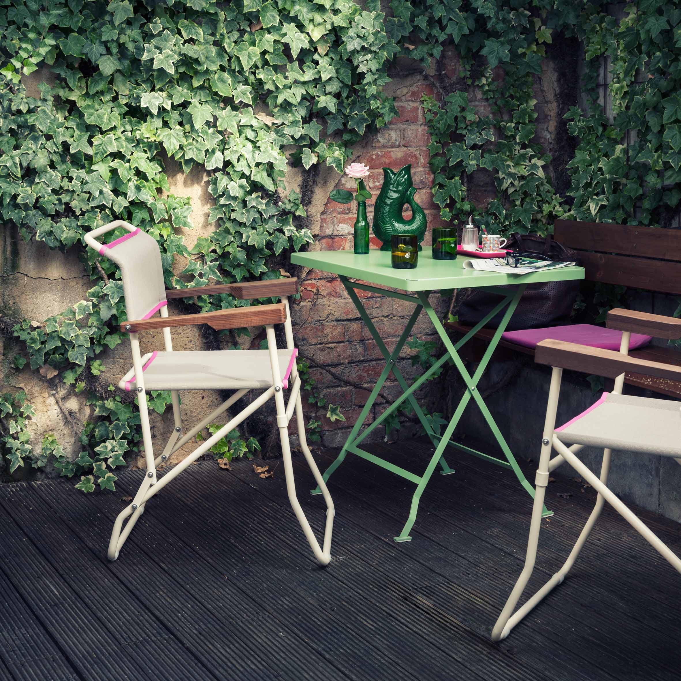 A cozy outdoor patio features two white chairs with wooden armrests around a Richard Lampert Flip Bistro Table, showcasing a green vase, lamp, and teacup. Lush ivy climbs a brick wall above an elegant wooden floor in the background.