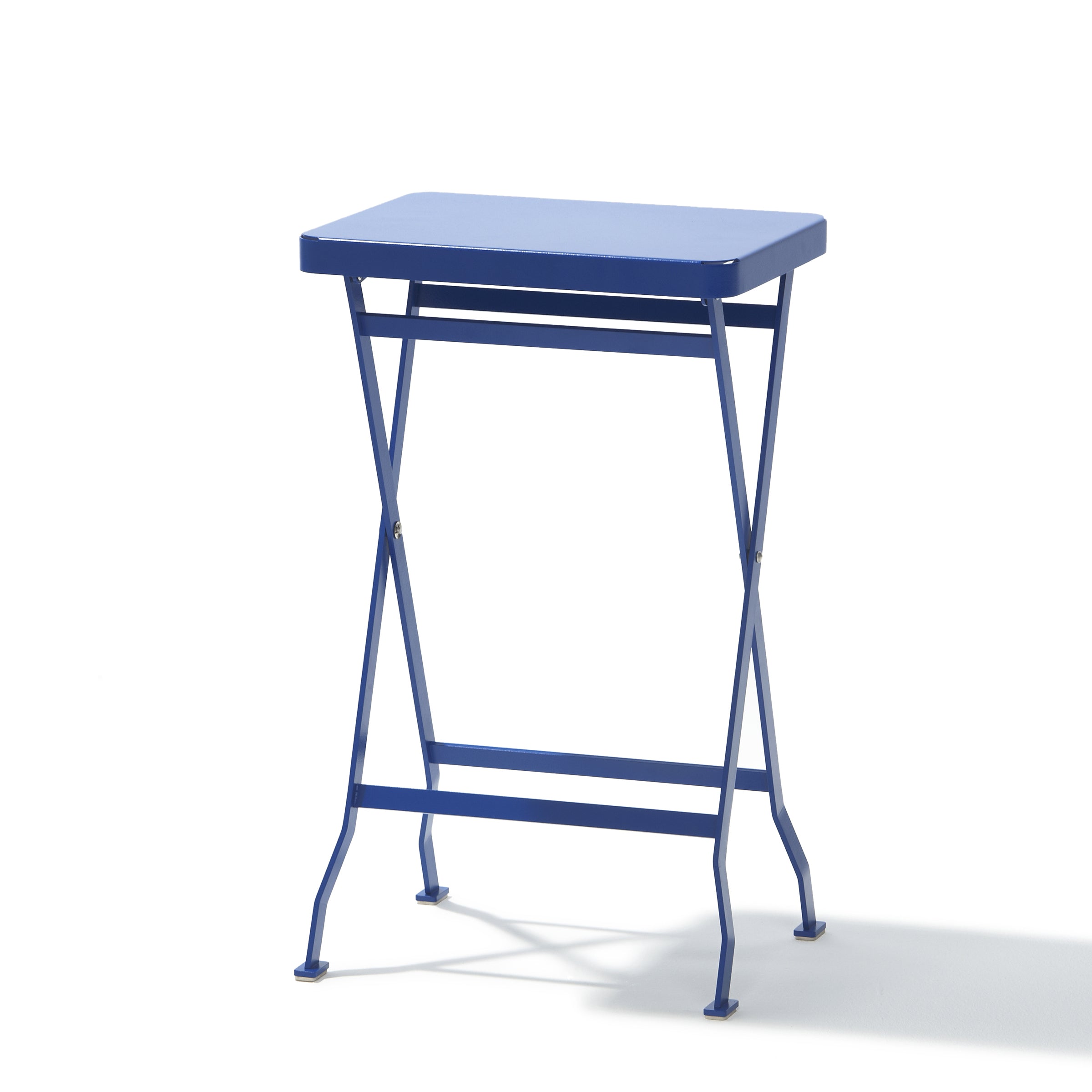 The Flip Side Table by Richard Lampert is a small, high-quality blue folding table with a rectangular top and slender crisscross legs. It stands on a white background, with shadows enhancing the bright lighting.