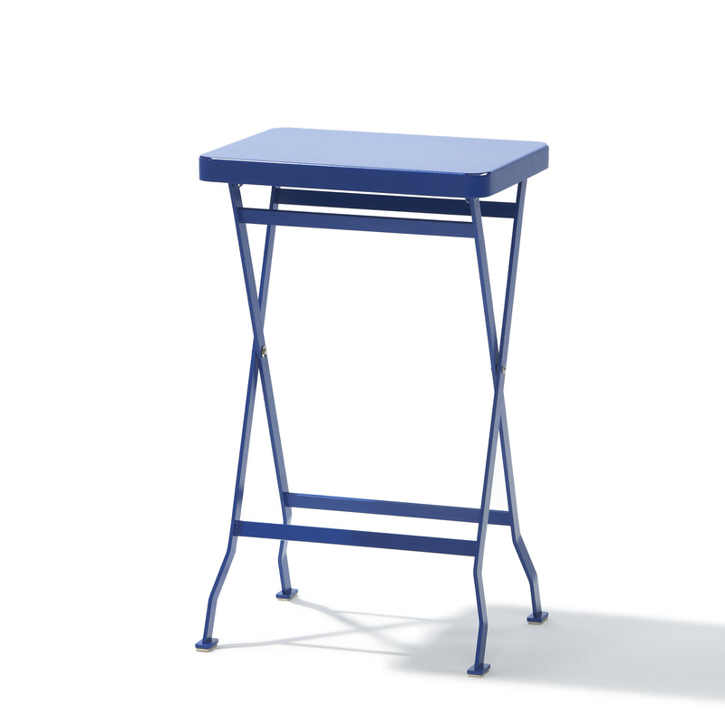 The Flip Side Table by Richard Lampert is a small, high-quality blue folding table with a rectangular top and slender crisscross legs. It stands on a white background, with shadows enhancing the bright lighting.