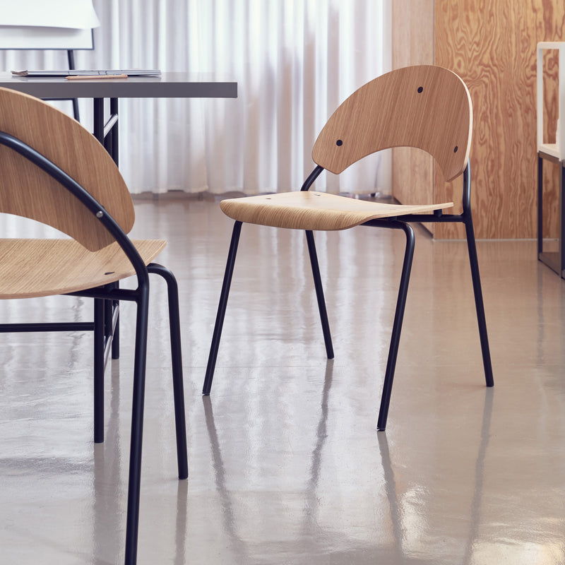 The Frog Chair by Richard Lampert, featuring a modern wooden design with a curved backrest and black metal legs reminiscent of Herbert Hirches style, is placed on a polished concrete floor. A table and another chair are partially visible amid a minimalist interior with white curtains and wood paneling.