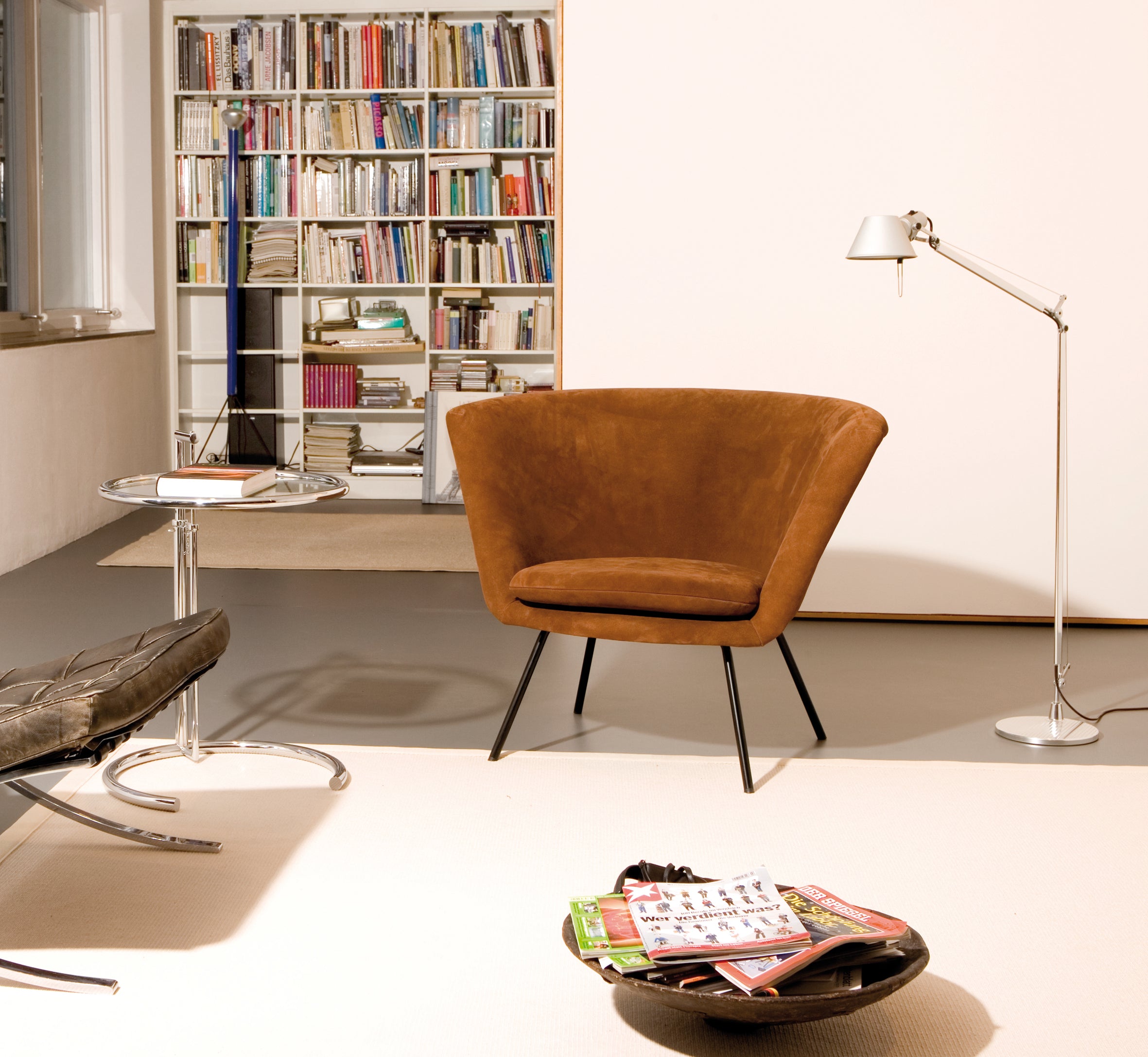A cozy room showcases Richard Lamperts H 57 Lounge Chair with a modern floor lamp, exuding mid-century design. A round side table holds a book, while a bookshelf brimming with reads is in the background. In the foreground, alongside a stainless steel chair, rests a bowl of magazines.