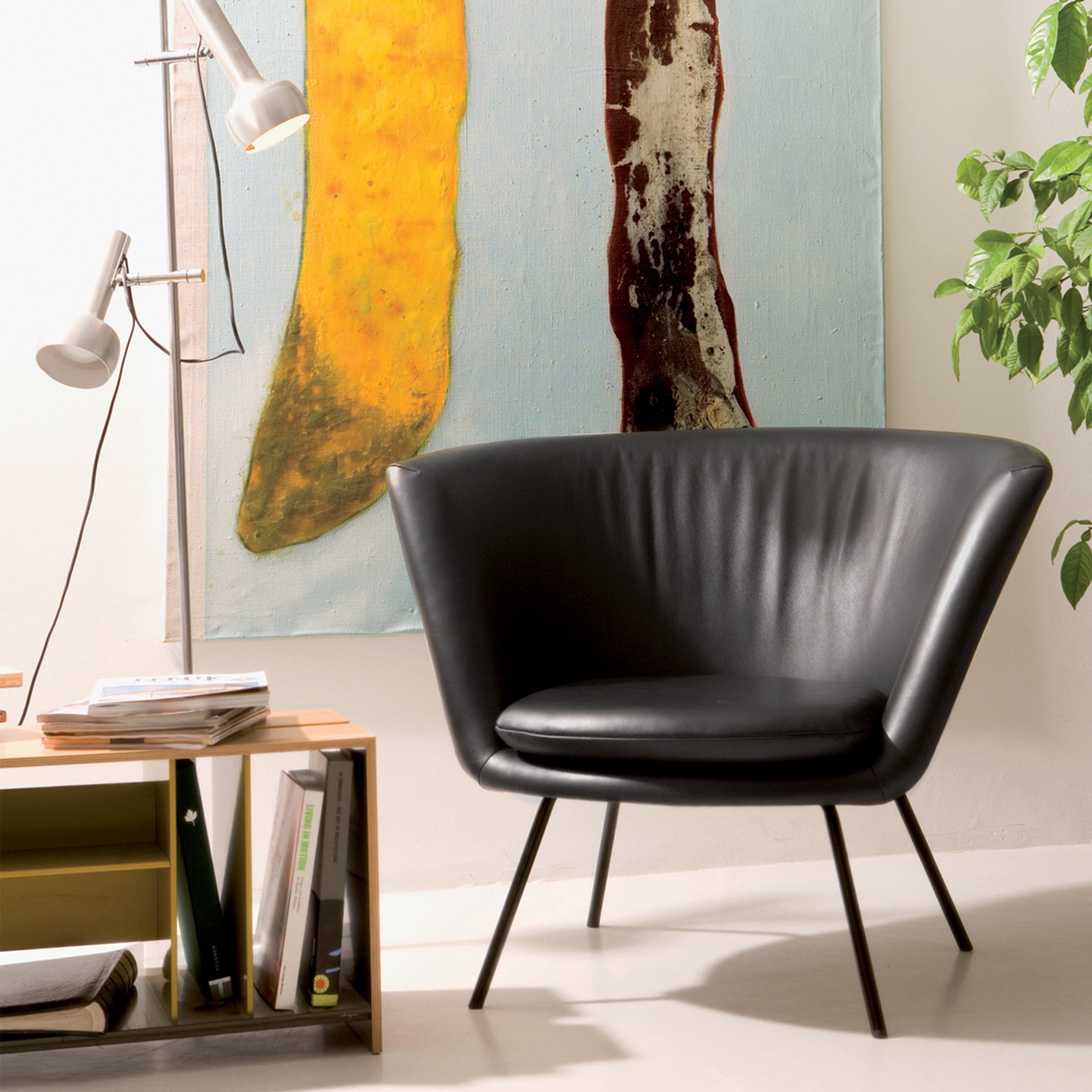 A modern room showcases the H 57 Lounge Chair by Richard Lampert, a black leather armchair with slim metal legs echoing mid-century design, paired with a floor lamp and wooden side table stacked with books. A large abstract painting with yellow and brown verticals decorates the wall, complemented by a green plant.
