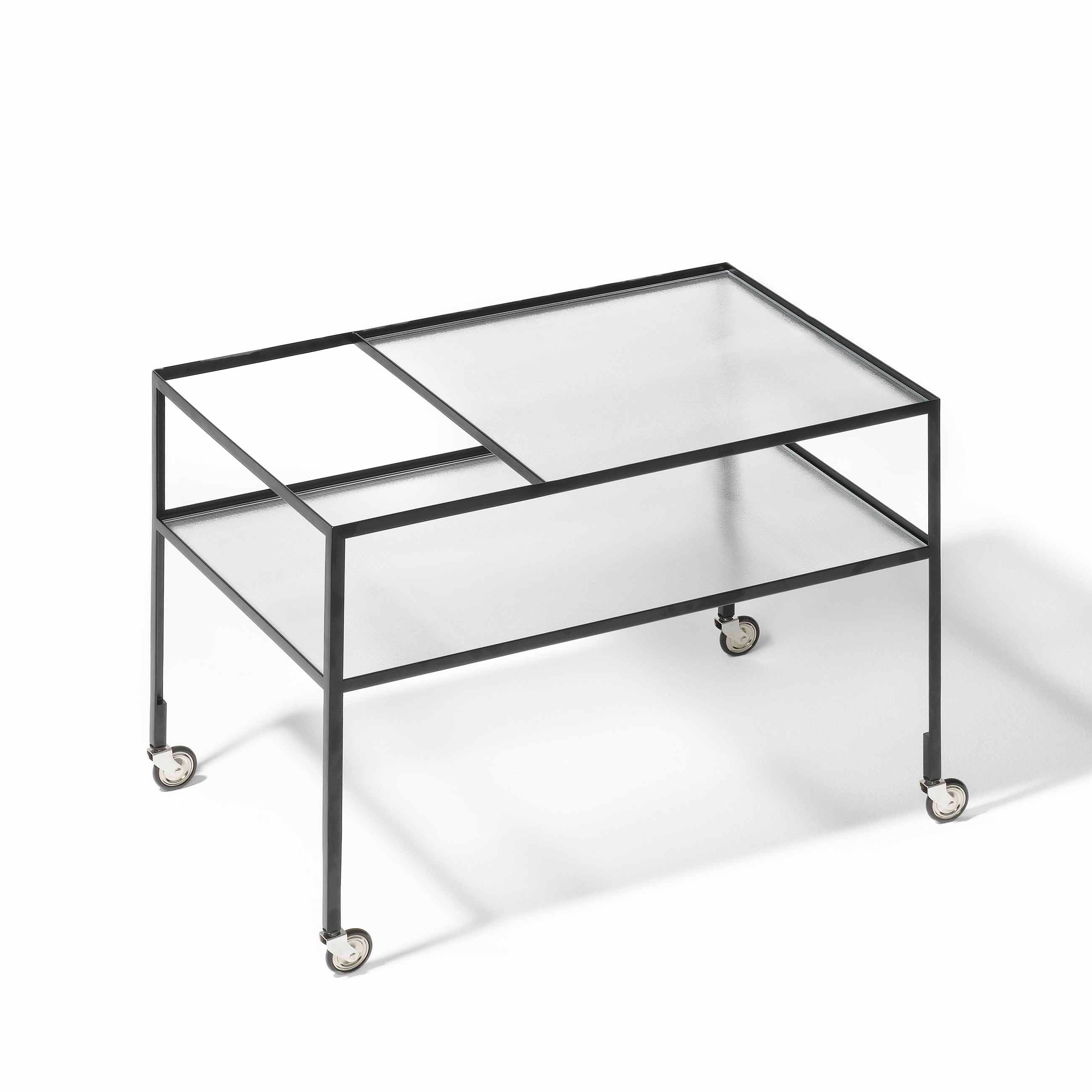 The Richard Lampert Bar Trolley showcases a minimalist design with a black steel frame, structured security glass top, open shelves, and four rolling wheels, exuding a modern industrial appeal.