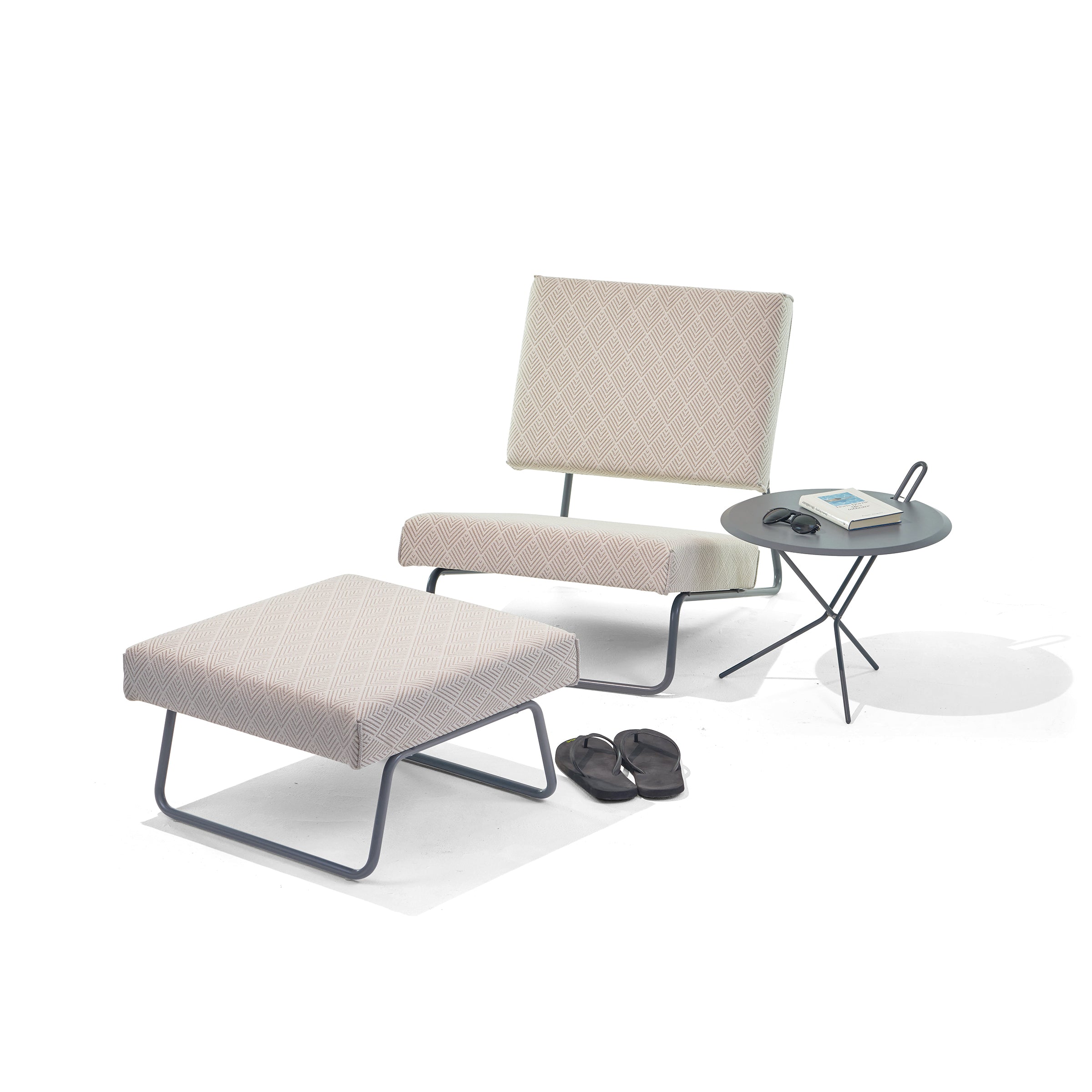 The Hirche Lounge Ottoman by Richard Lampert features a modern Bauhaus design with a light geometric pattern. A small round side table holds a book and eyeglasses, while black slippers rest nearby, showcasing the timeless elegance inspired by Herbert Hirche.