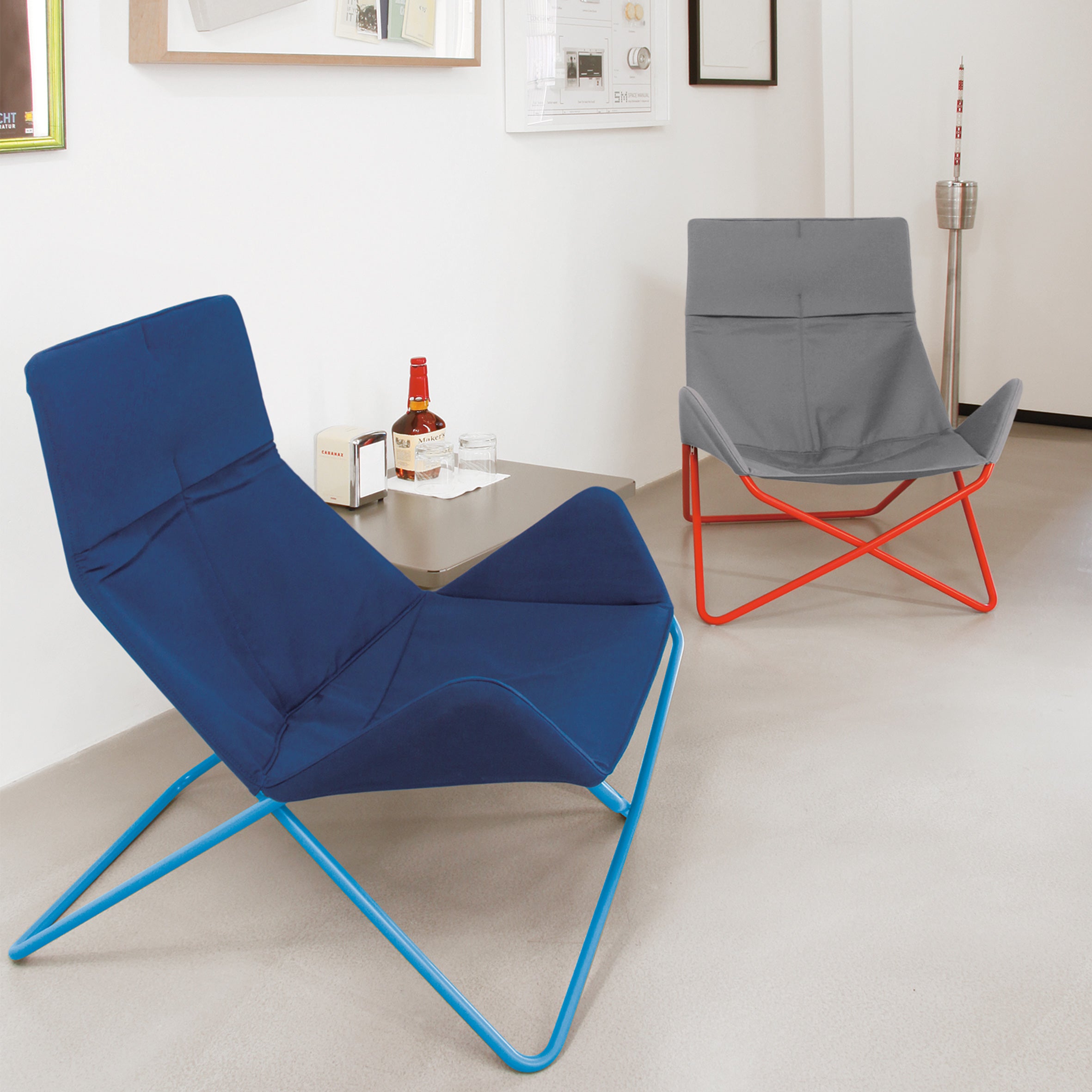 Two Richard Lampert In-Out Chairs—one with a blue frame and seat, the other with a red frame and gray seat—are on a light floor. A small metal table between them holds items, including a bottle, in this minimalist outdoor room.