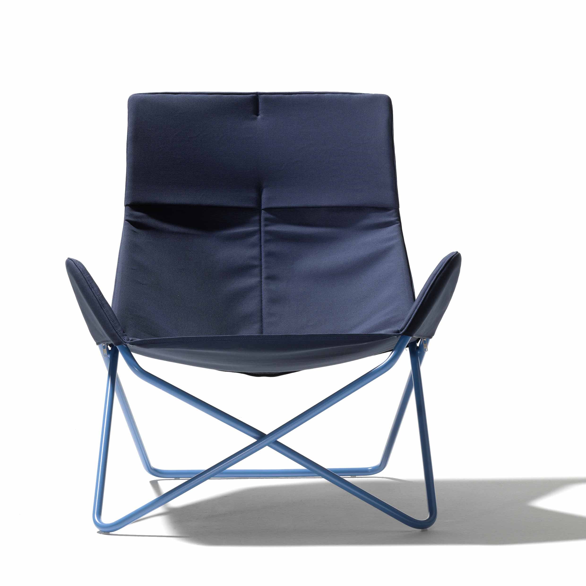The In-Out Chair by Richard Lampert is a modern, lightweight armchair with a navy blue fabric seat and minimalist design. It has a weather-resistant metal frame and crisscross leg structure, casting a shadow on white.