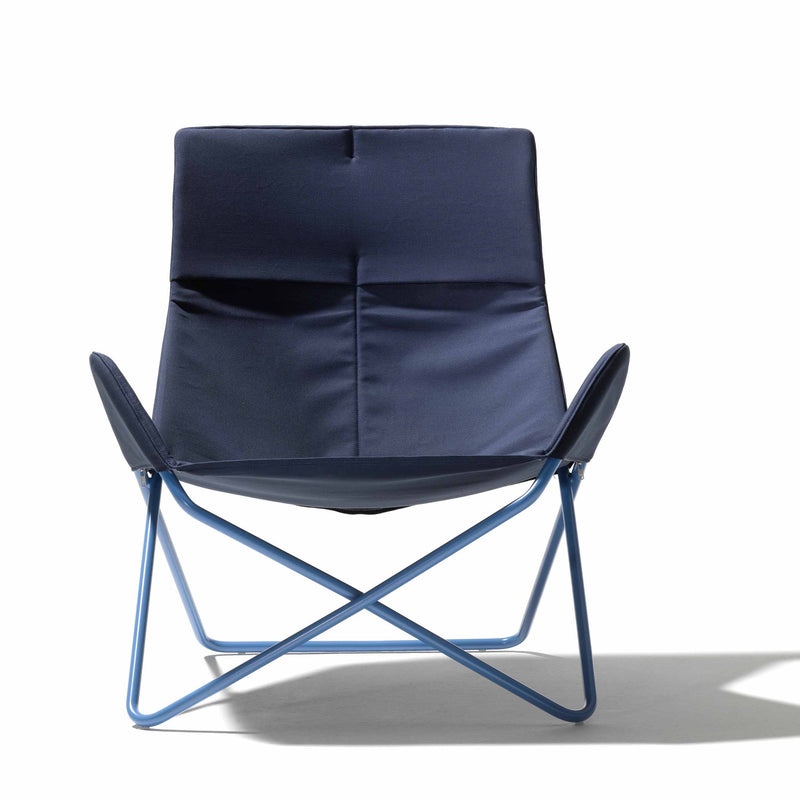 The In-Out Chair by Richard Lampert is a modern, lightweight armchair with a navy blue fabric seat and minimalist design. It has a weather-resistant metal frame and crisscross leg structure, casting a shadow on white.